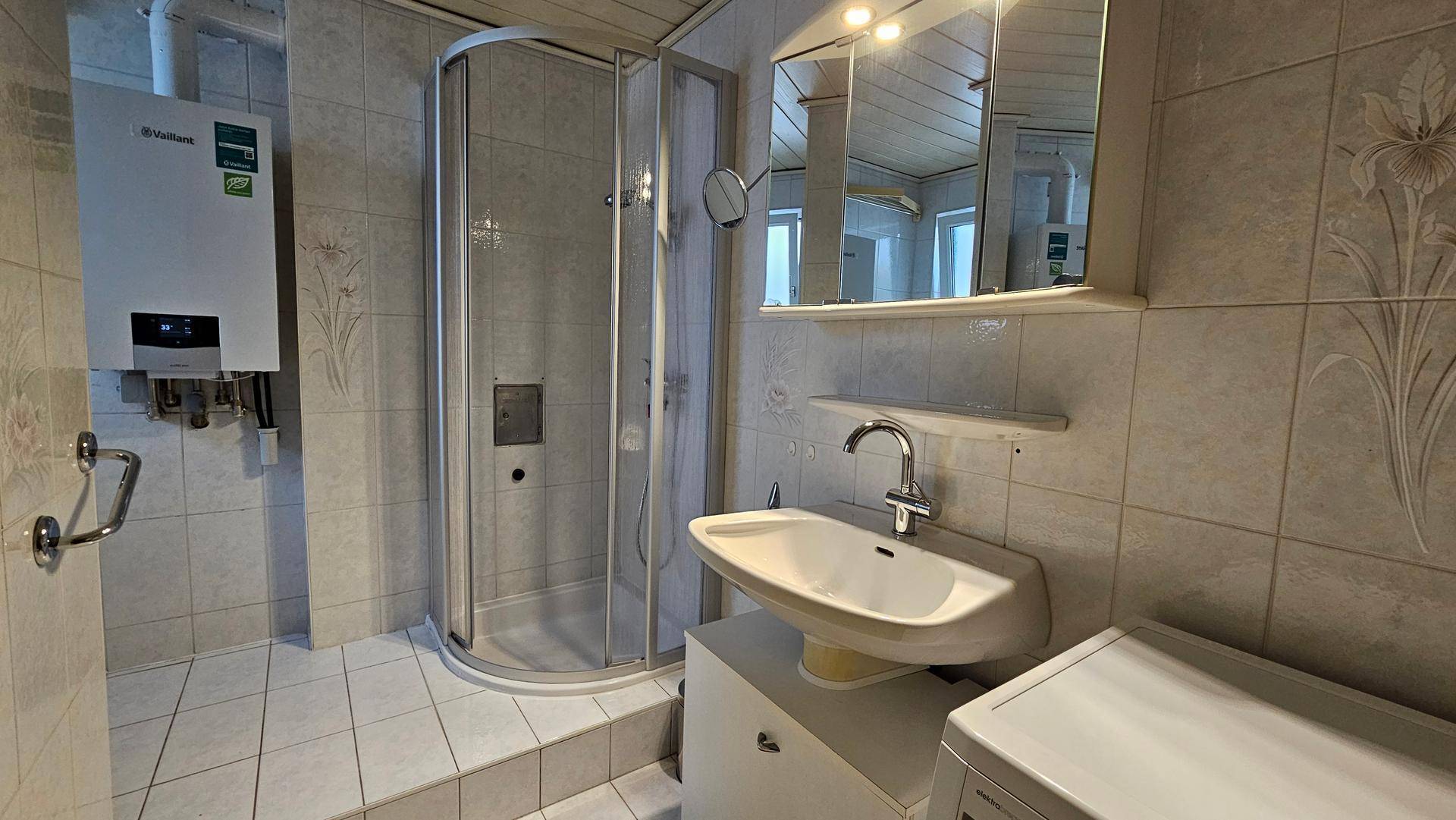 Badezimmer mit Duschkabine, Waschbecken und Waschmaschine, hell gefliest.