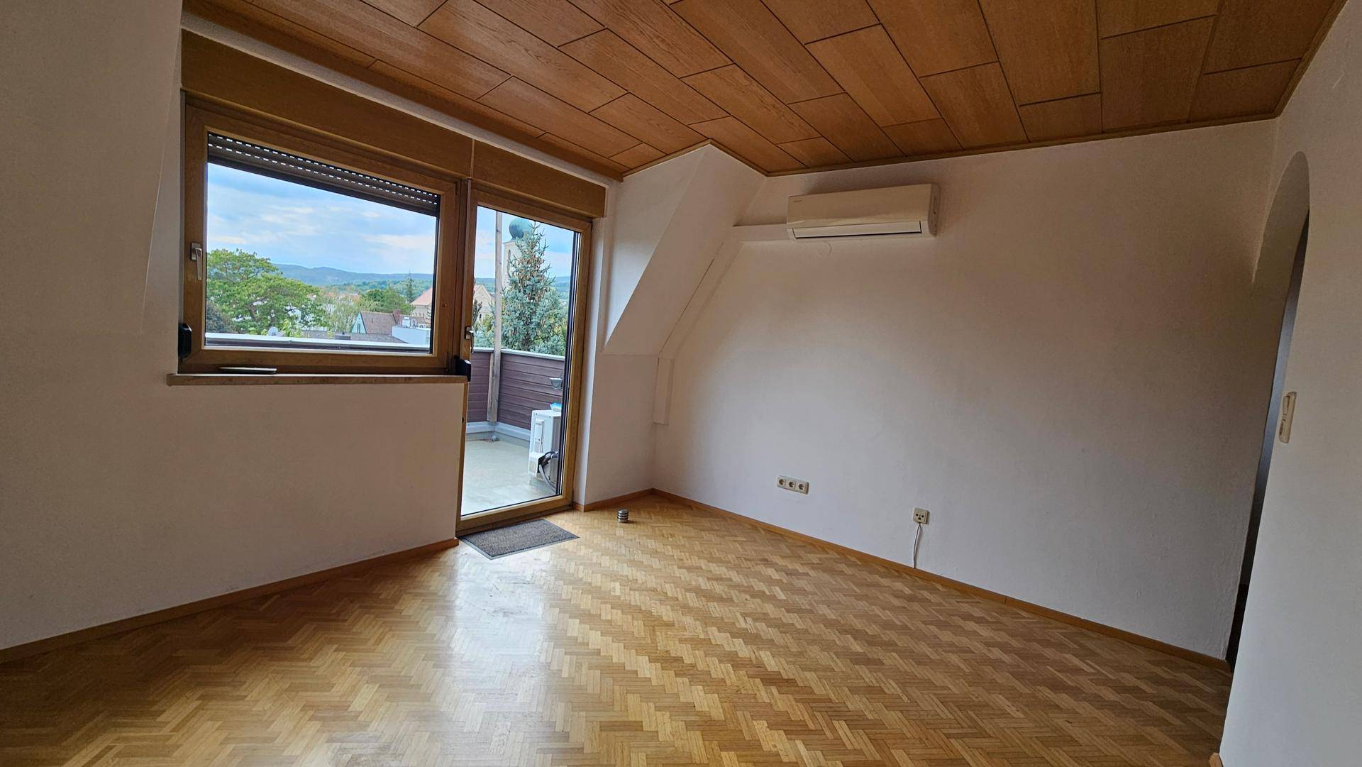 Wohnbereich mit Parkettboden, Klimaanlage und großem Fenster mit Blick ins Grüne.