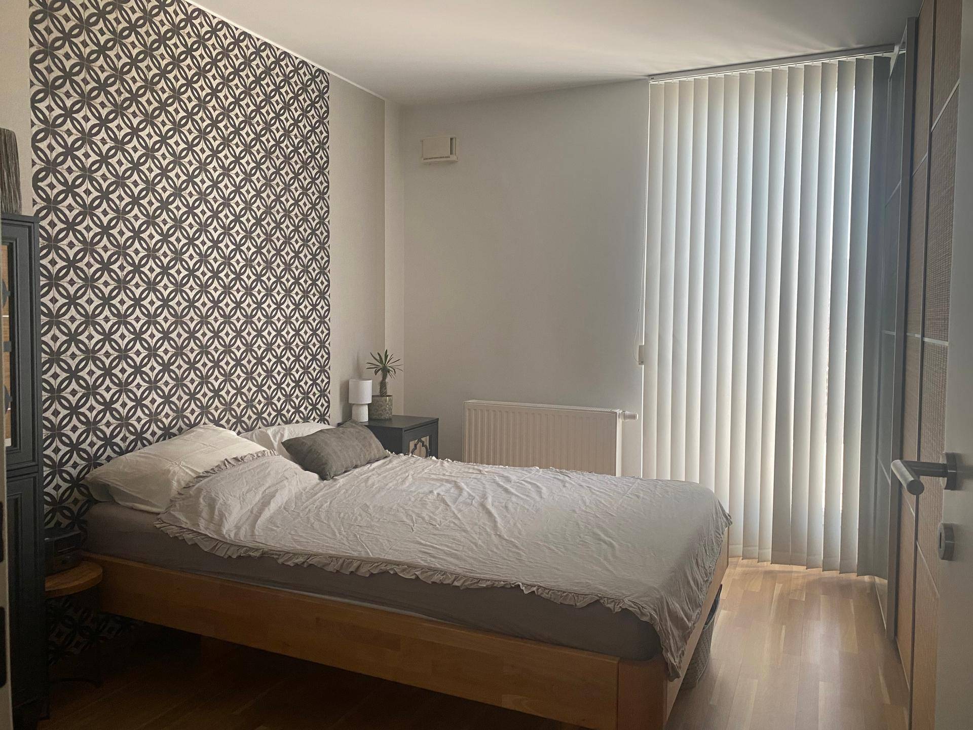 Schlafzimmer mit Holzbett, gemusterter Akzentwand und Fenster mit vertikalen Jalousien.