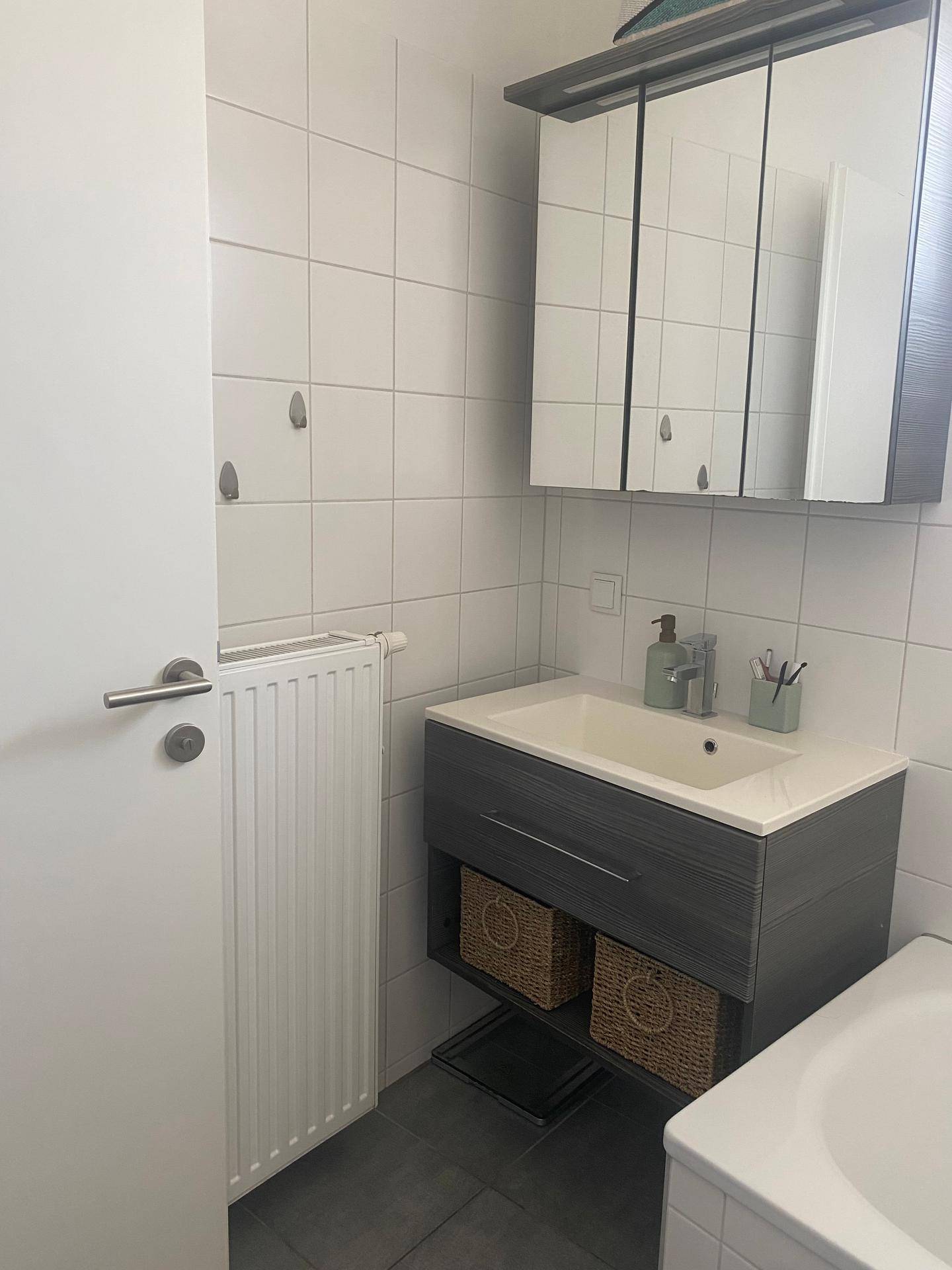 Badezimmer mit weißen Fliesen, grauem Waschtisch, Spiegelschrank und Heizkörper.