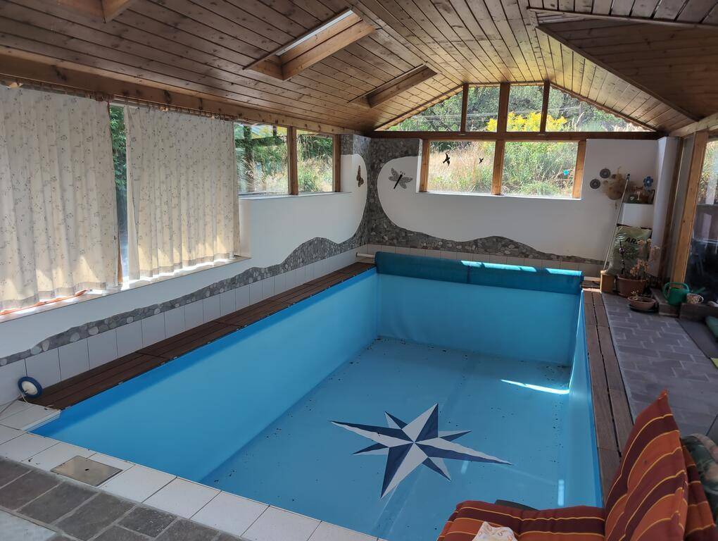 Innenbereich des Poolhauses mit Schwimmbecken, Holzdecke und großen Fenstern, die viel Licht hereinlassen.