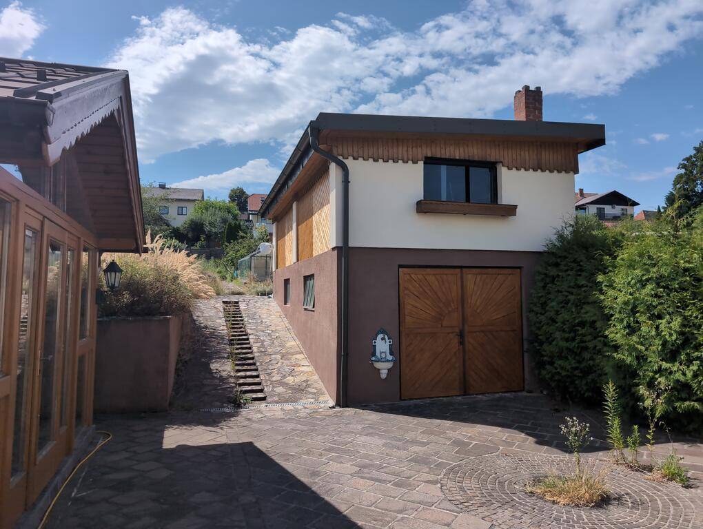 Seitenansicht des Hauses mit weiterer Garage, Außentreppe und gepflastertem Hof unter blauem Himmel.