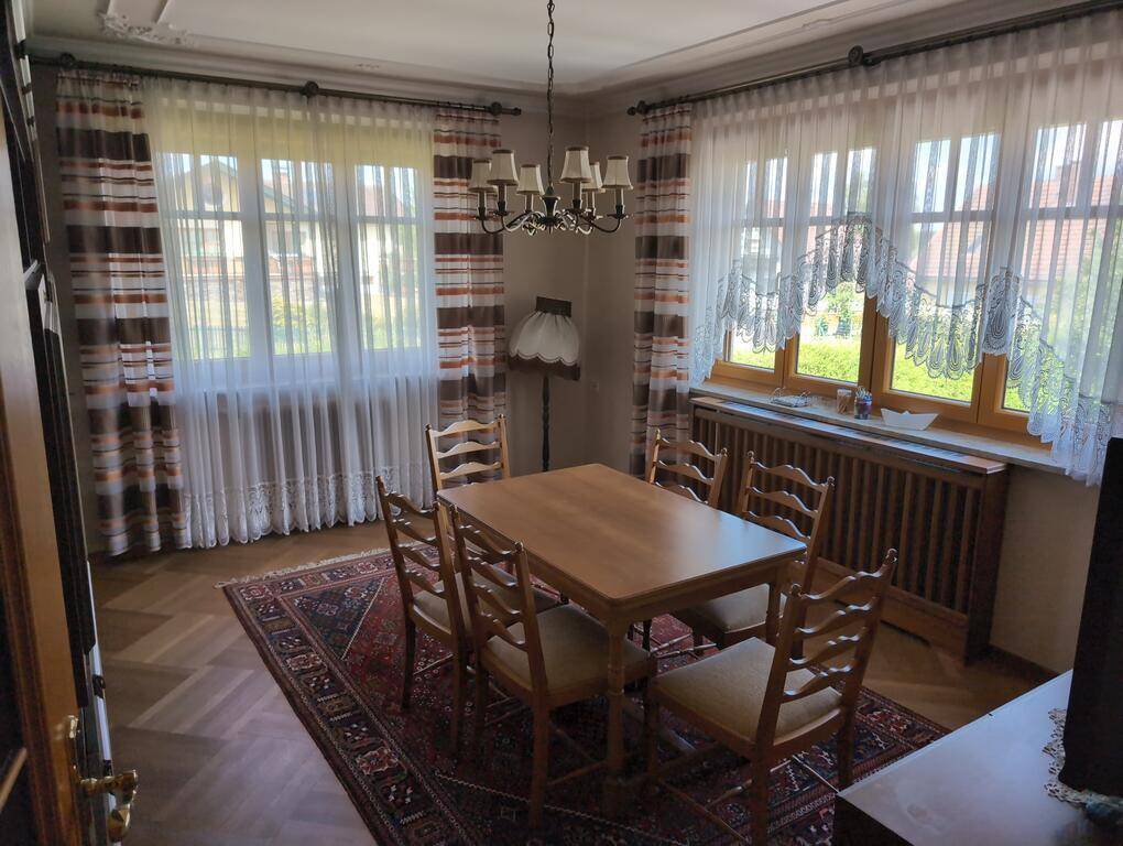 Esszimmer mit Holztisch, Stühlen, Parkettboden und großen Fenstern, die eine helle Atmosphäre schaffen.