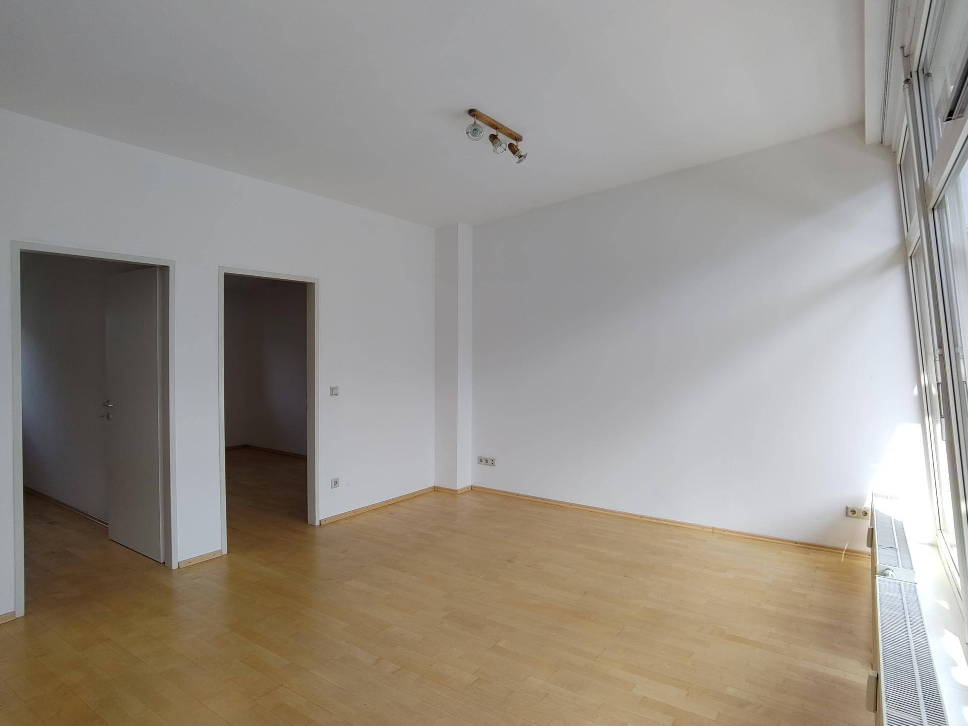Helles, leeres Wohnzimmer mit Parkettboden und großen Fenstern.
