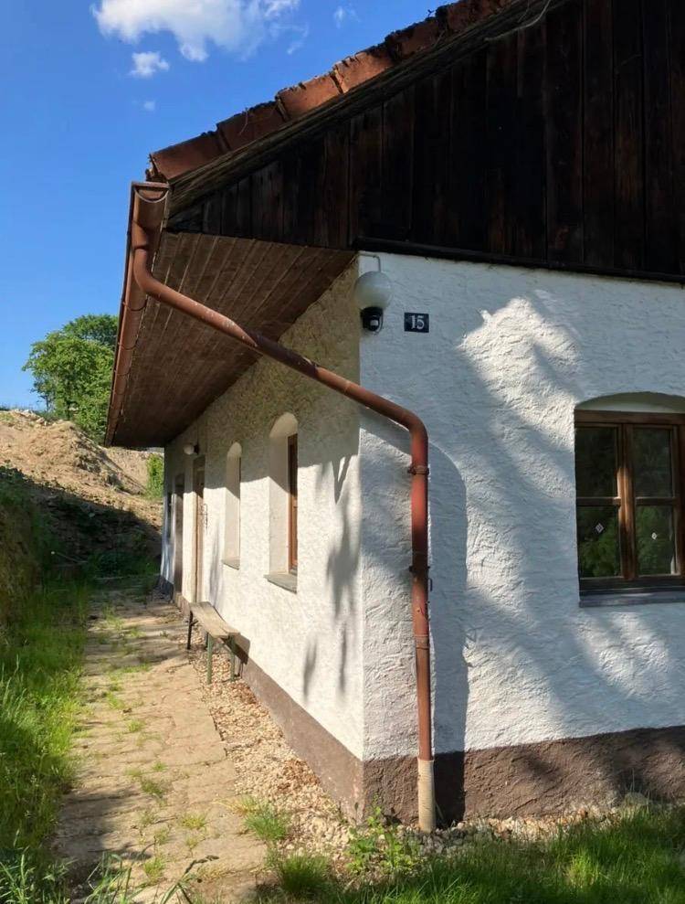 Weiße Fassade des Hauses mit braunen Fensterrahmen und einer Regenrinne, umgeben von einfacher Vegetation.