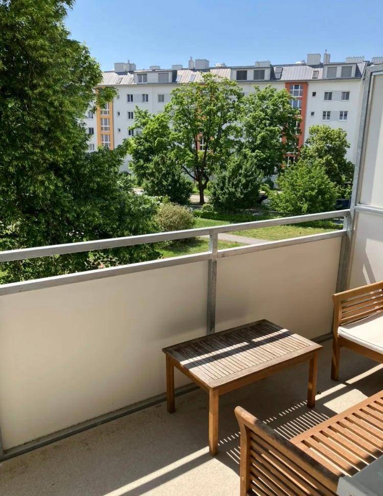 Möblierter Balkon mit Holzsitzgelegenheiten und Blick auf grüne Bäume und Wohngebäude.