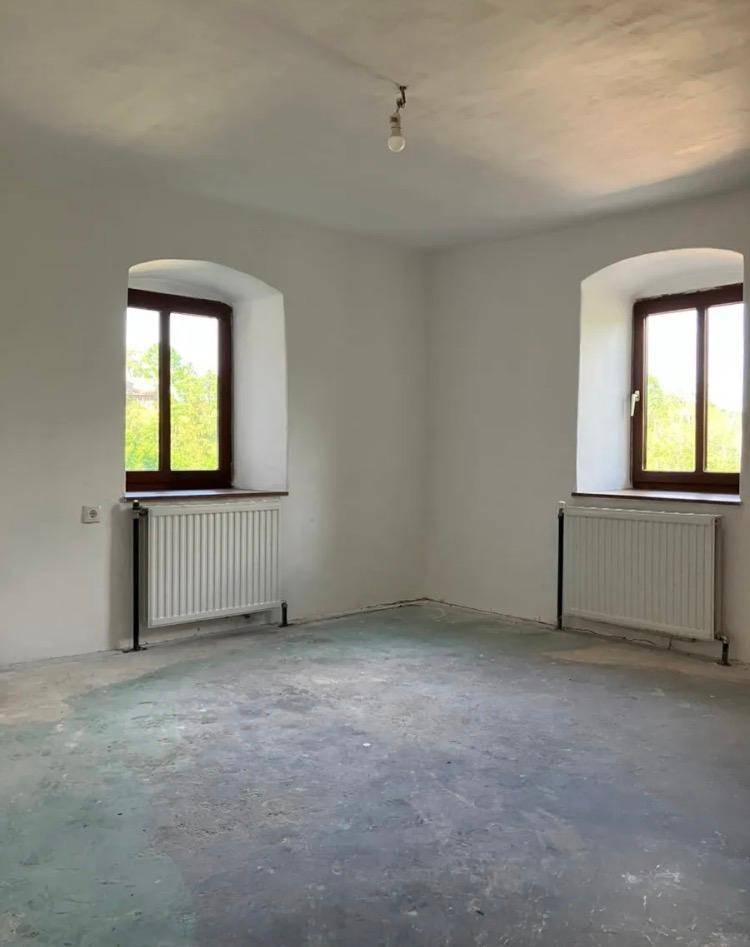 Leerer Raum mit zwei Fenstern, Heizkörpern und einem unfertigen Betonboden, bereit zur Gestaltung.