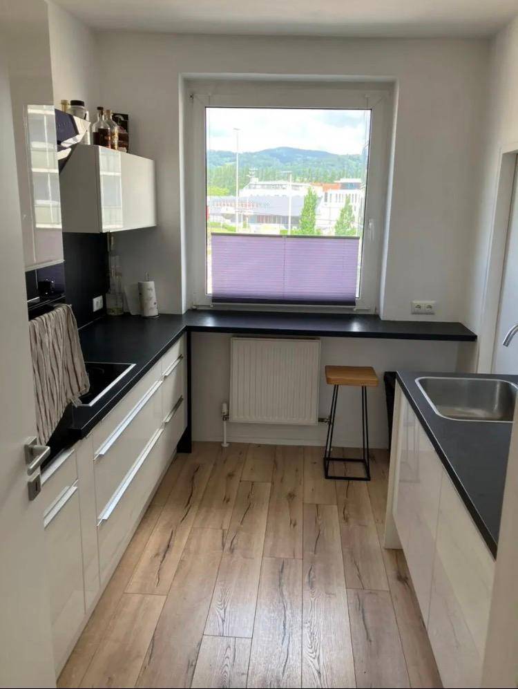 Helle Küche mit Fenster, Holzfußboden und schwarzer Arbeitsplatte, die einen Blick ins Grüne bietet.