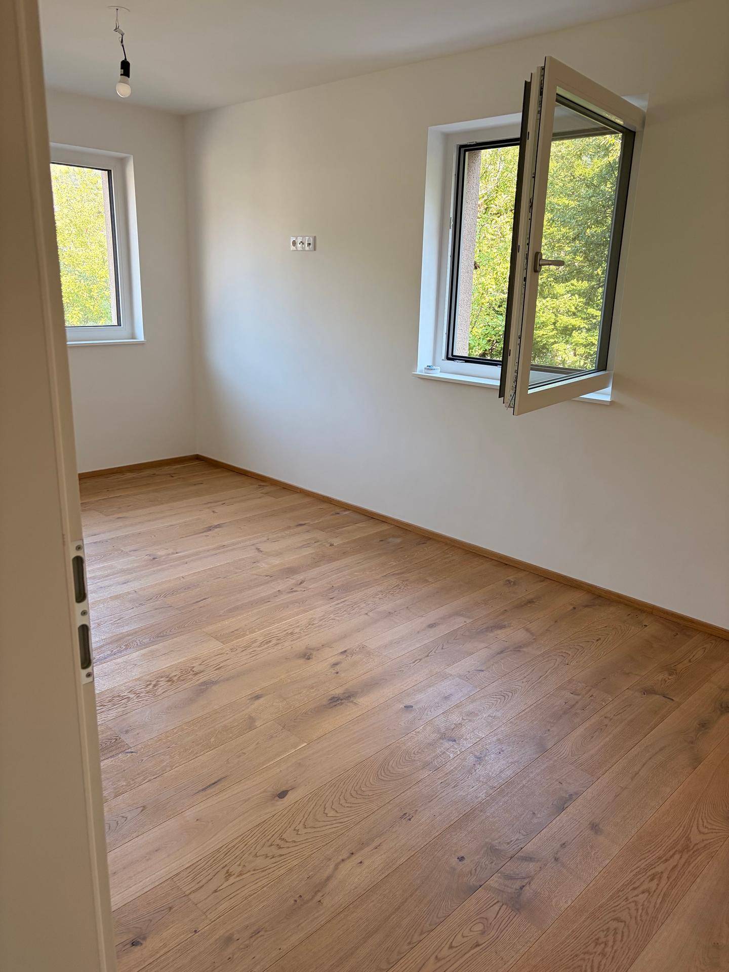 Helles Schlafzimmer mit Holzboden und zwei Fenstern mit Blick ins Grüne.