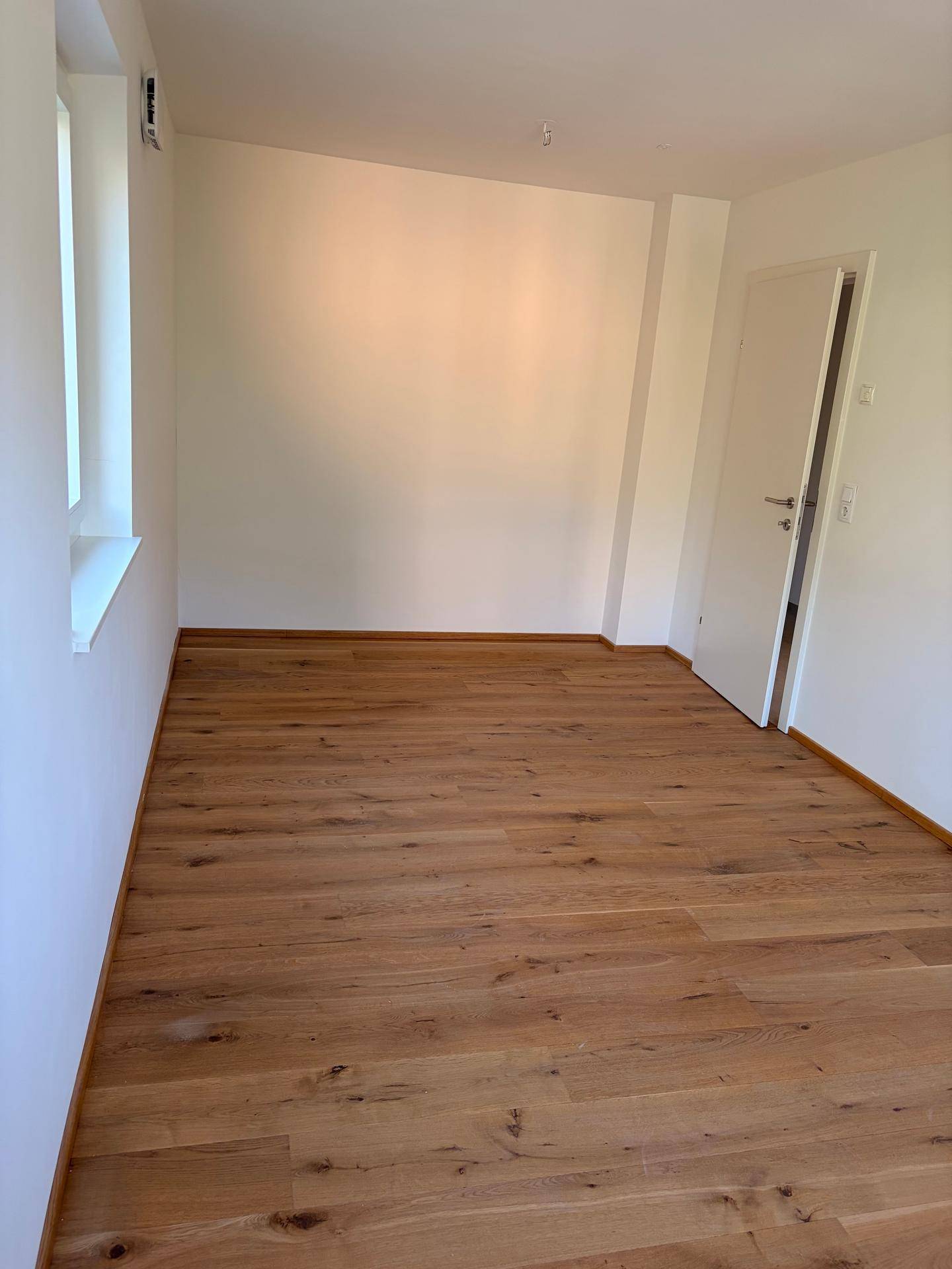 Geräumiges Zimmer mit Holzboden, weißen Wänden und viel Tageslicht.