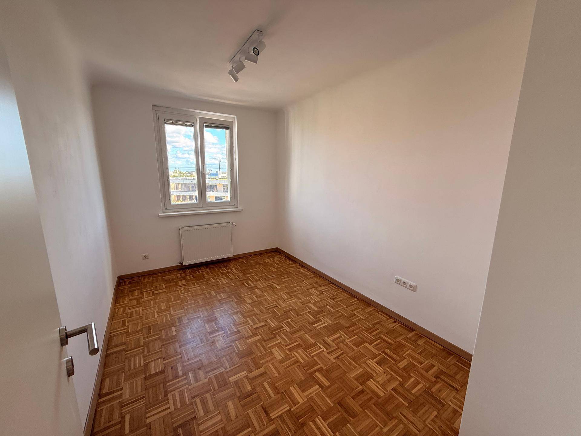 Helles Zimmer mit Parkettboden und einem Fenster, das viel Tageslicht hereinlässt.