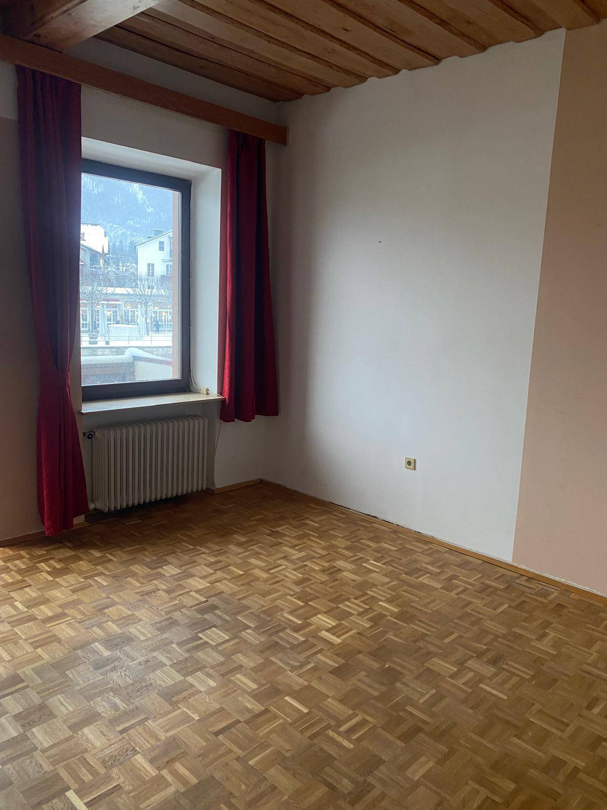 Helles Zimmer mit Parkettboden, Holzbalkendecke und einem Fenster mit rotem Vorhang.