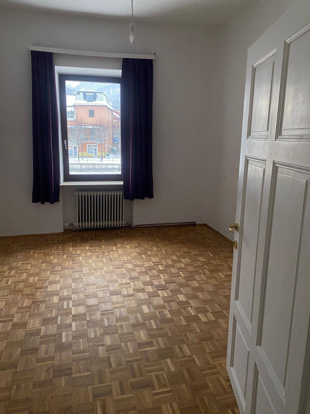 Leeres Zimmer mit Parkettboden und einem Fenster, das viel Tageslicht hereinlässt.