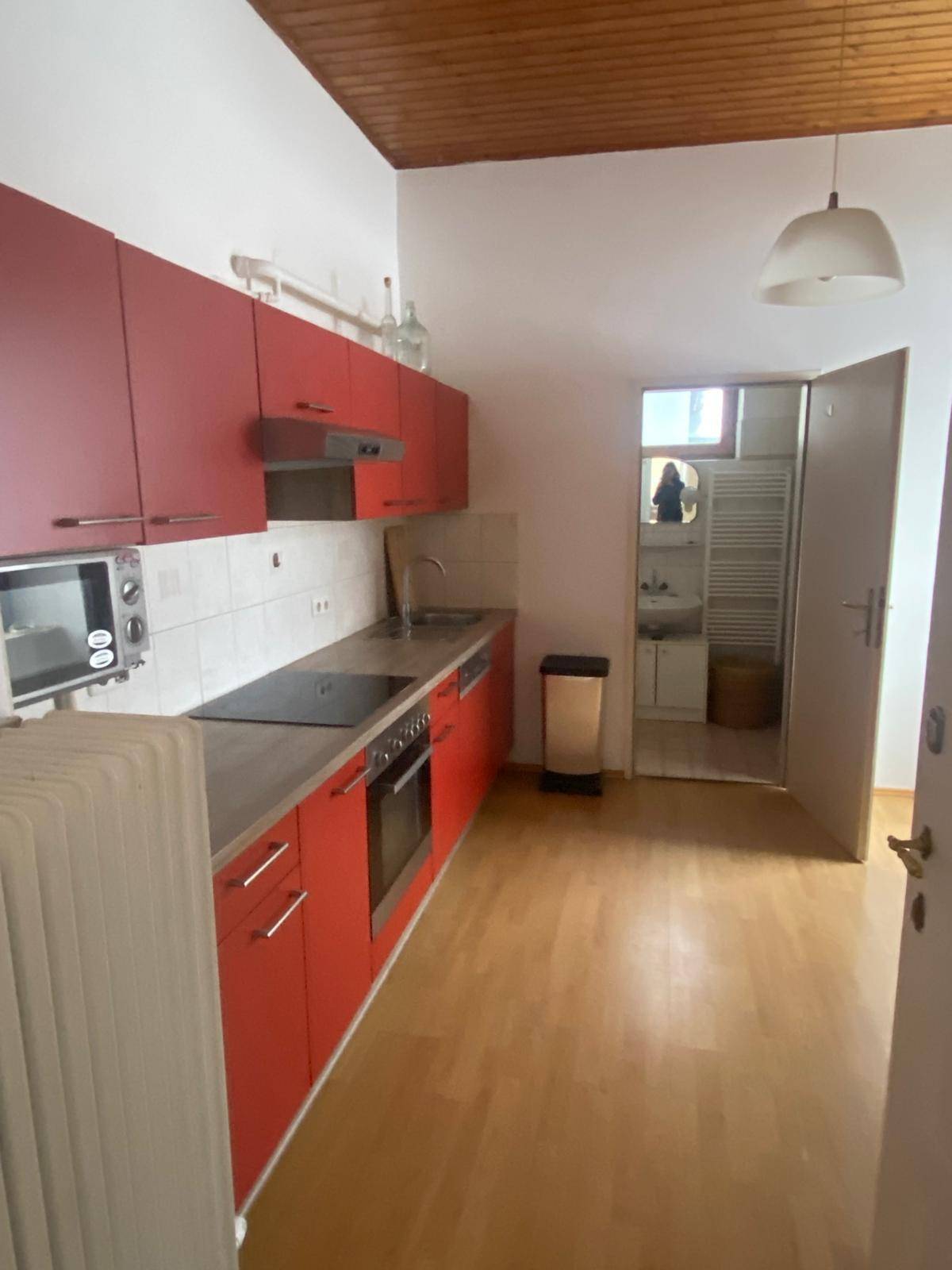 Funktionale Küche mit roten Schränken, Holzboden und Blick in ein angrenzendes Badezimmer.