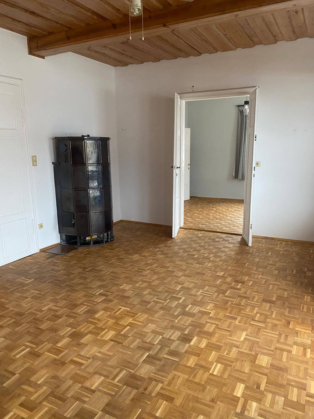 Helles Zimmer mit Parkettboden, Holzbalkendecke und einem traditionellen Kachelofen.