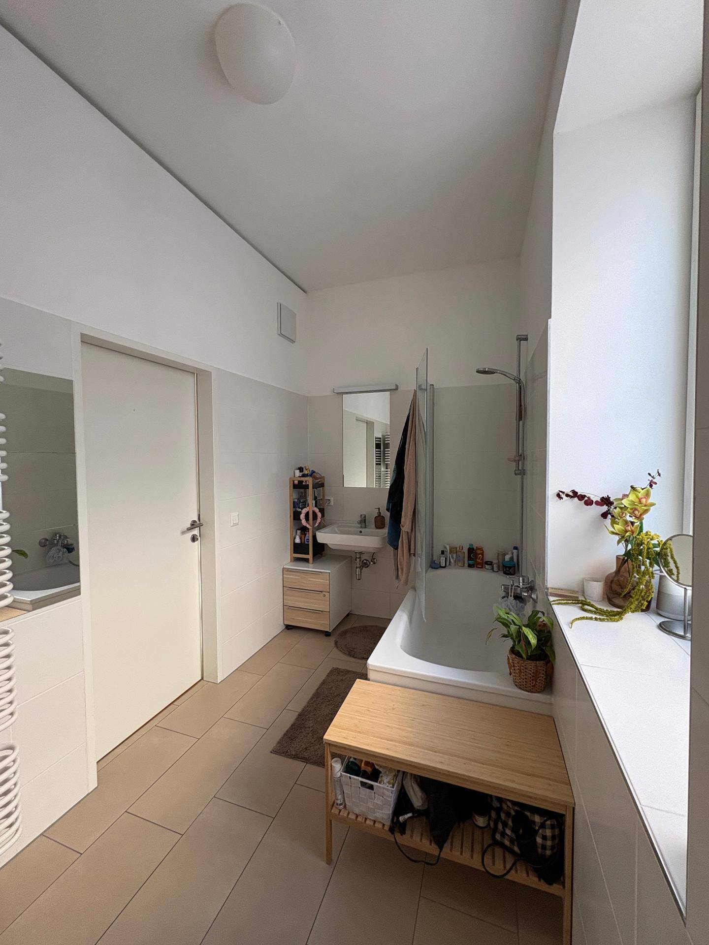 Badezimmer mit Badewanne, Waschbecken und Fenster, auf hellem Fliesenboden.