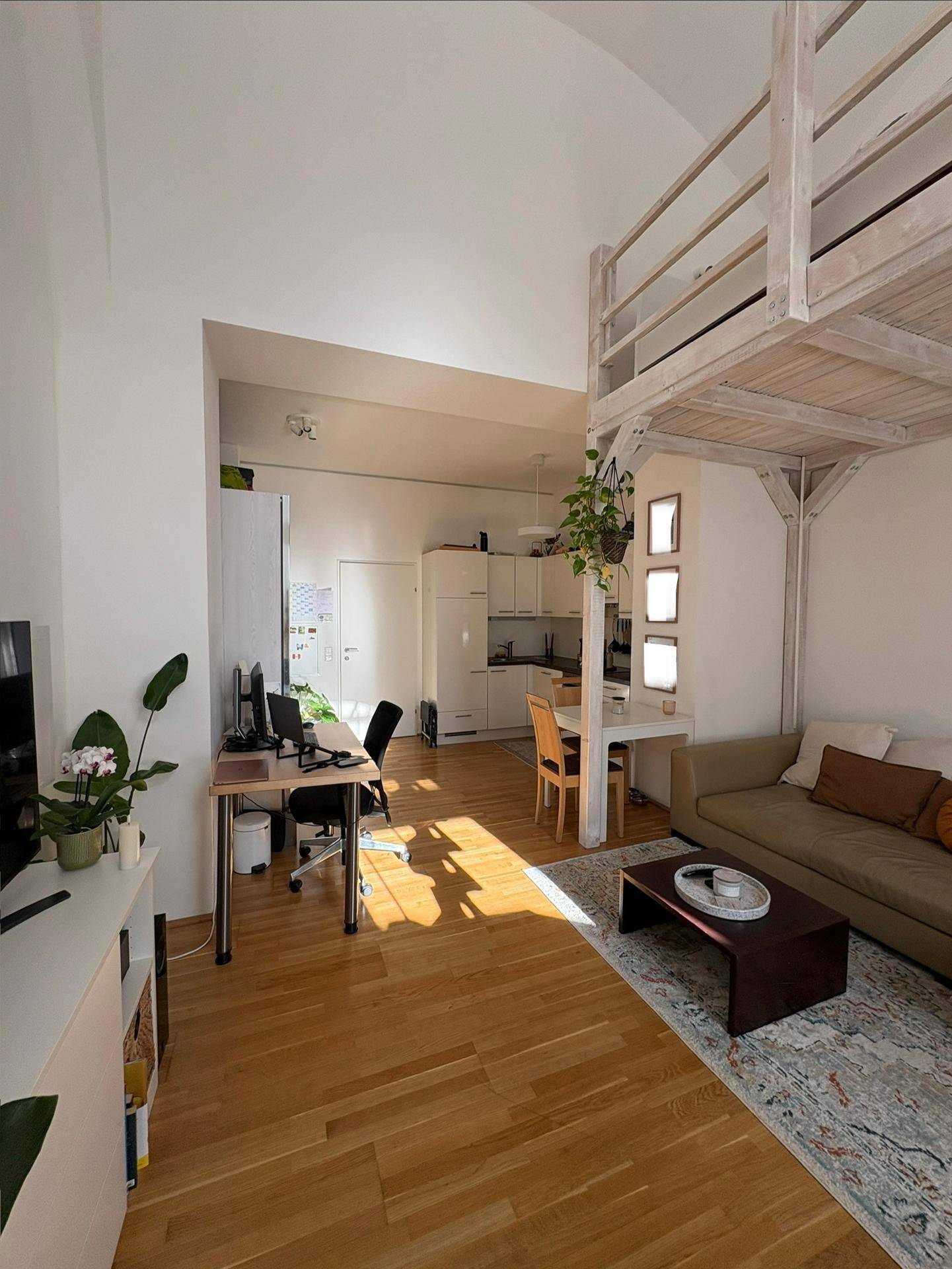 Heller Wohnbereich mit Arbeitsplatz, Sofa und offener Küche, ergänzt durch eine Loft-Struktur.