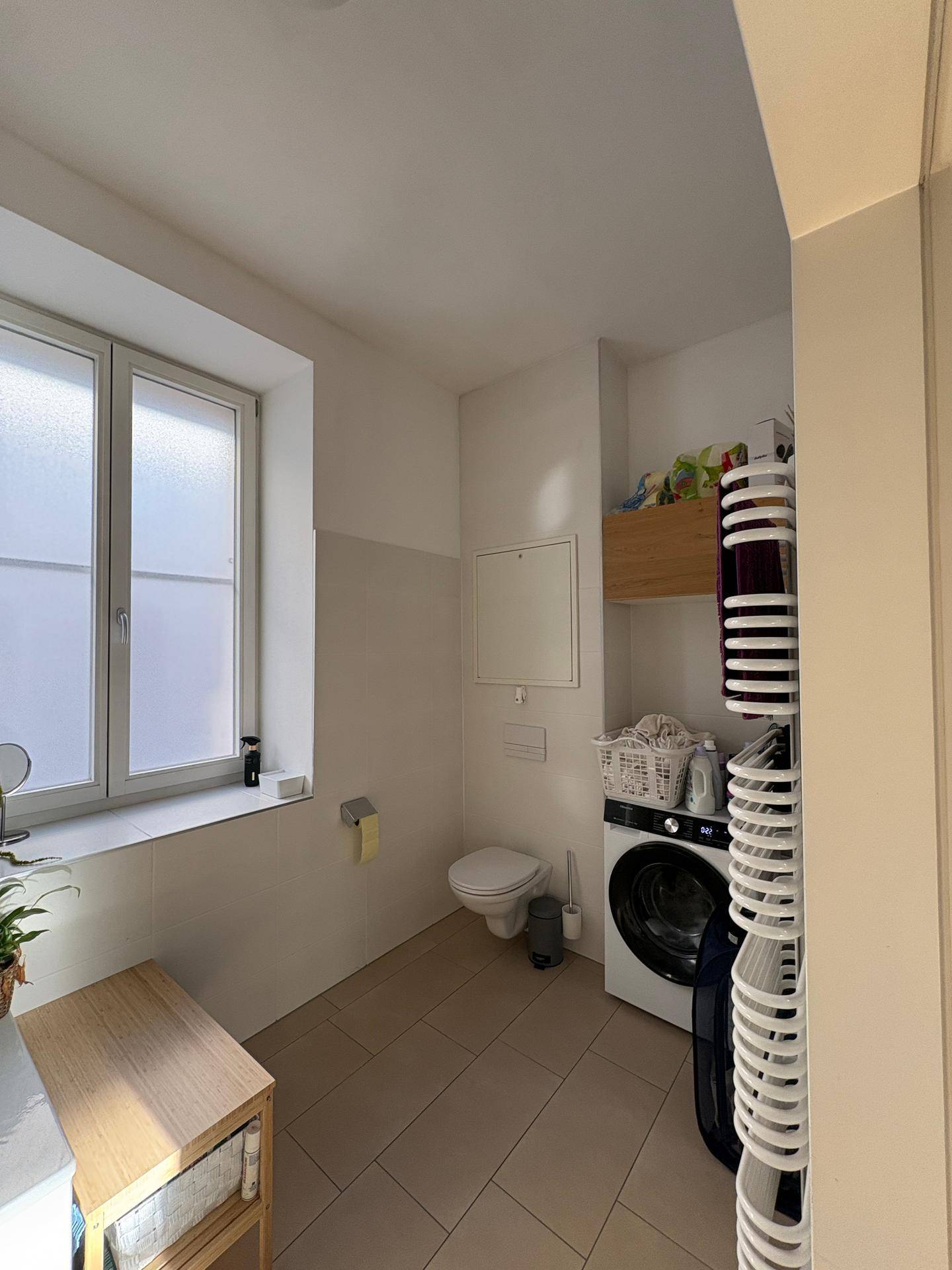 Helles Badezimmer mit Toilette, Waschmaschine und Fenster, ausgestattet mit Fliesenboden.