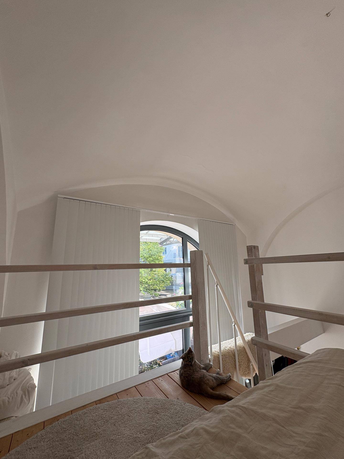 Blick vom Loft-Schlafzimmer auf das große Bogenfenster mit Jalousien und viel Tageslicht.