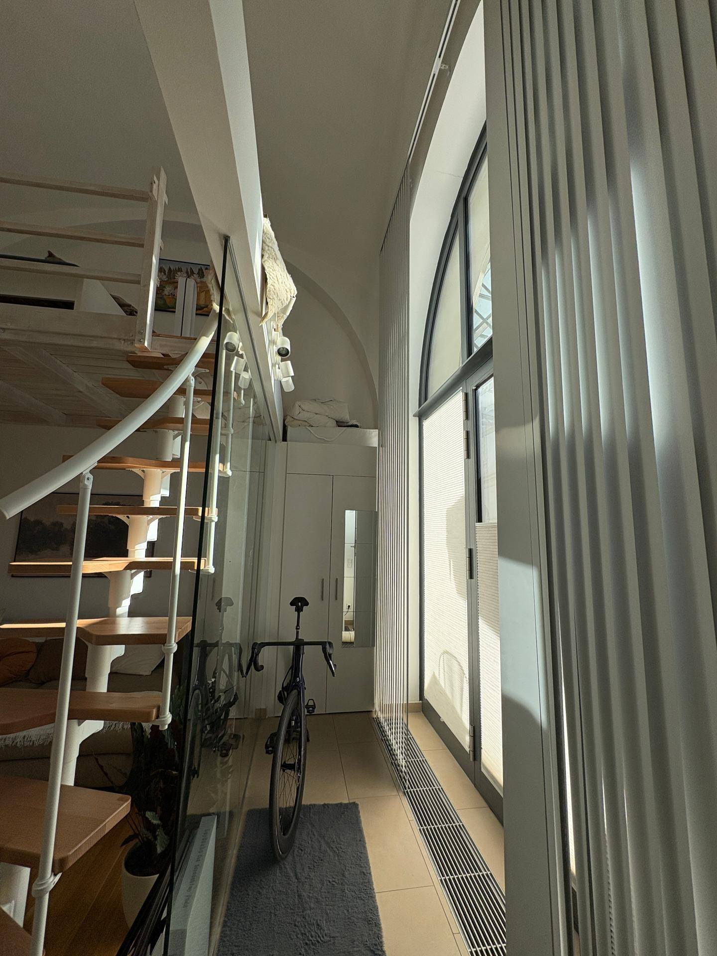 Wendeltreppe neben einem hohen Bogenfenster mit Jalousien, führt zum oberen Loftbereich.