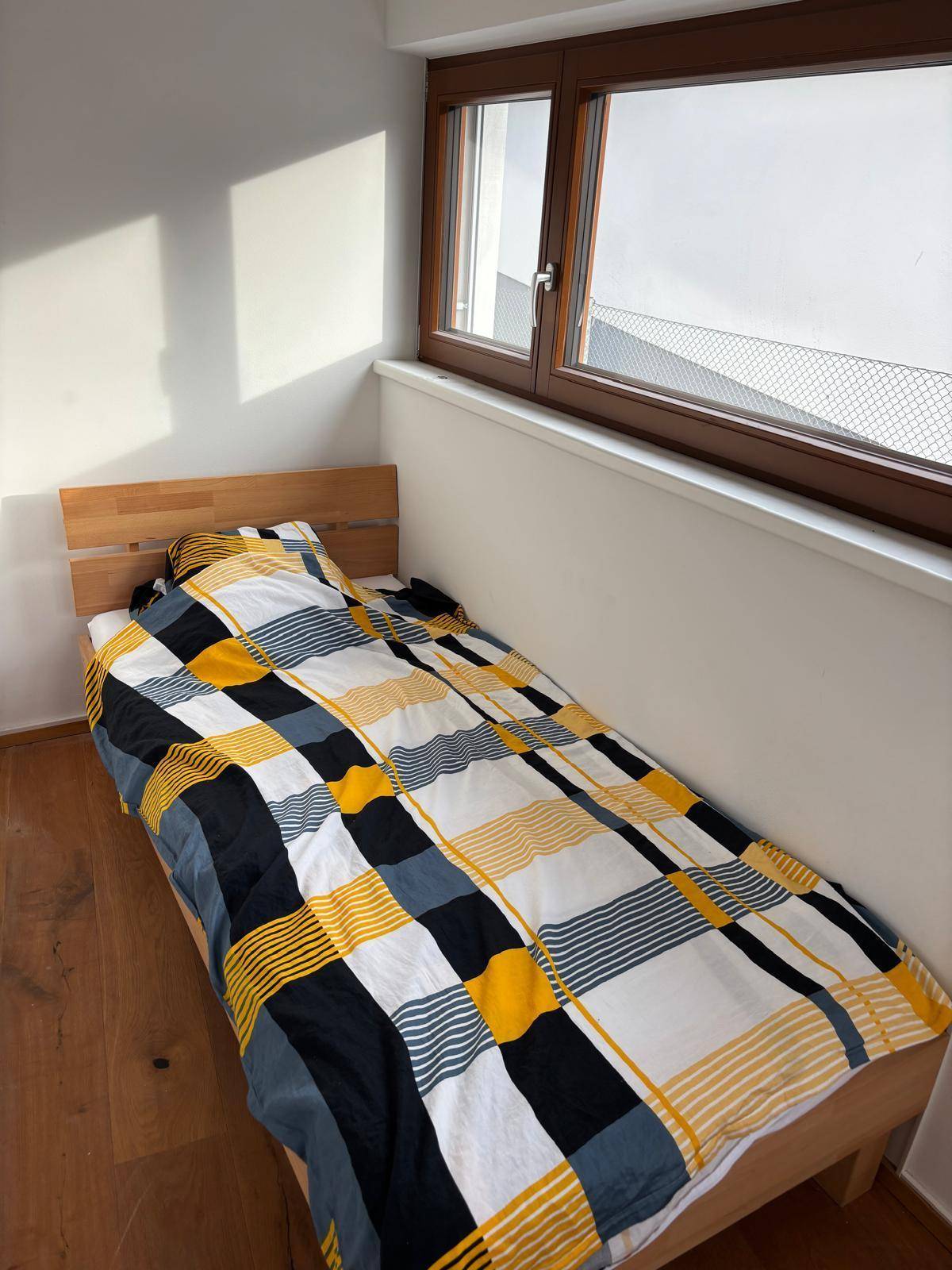 Schlafzimmer mit Einzelbett und farbenfroher Bettwäsche, beleuchtet durch ein großes Fenster.