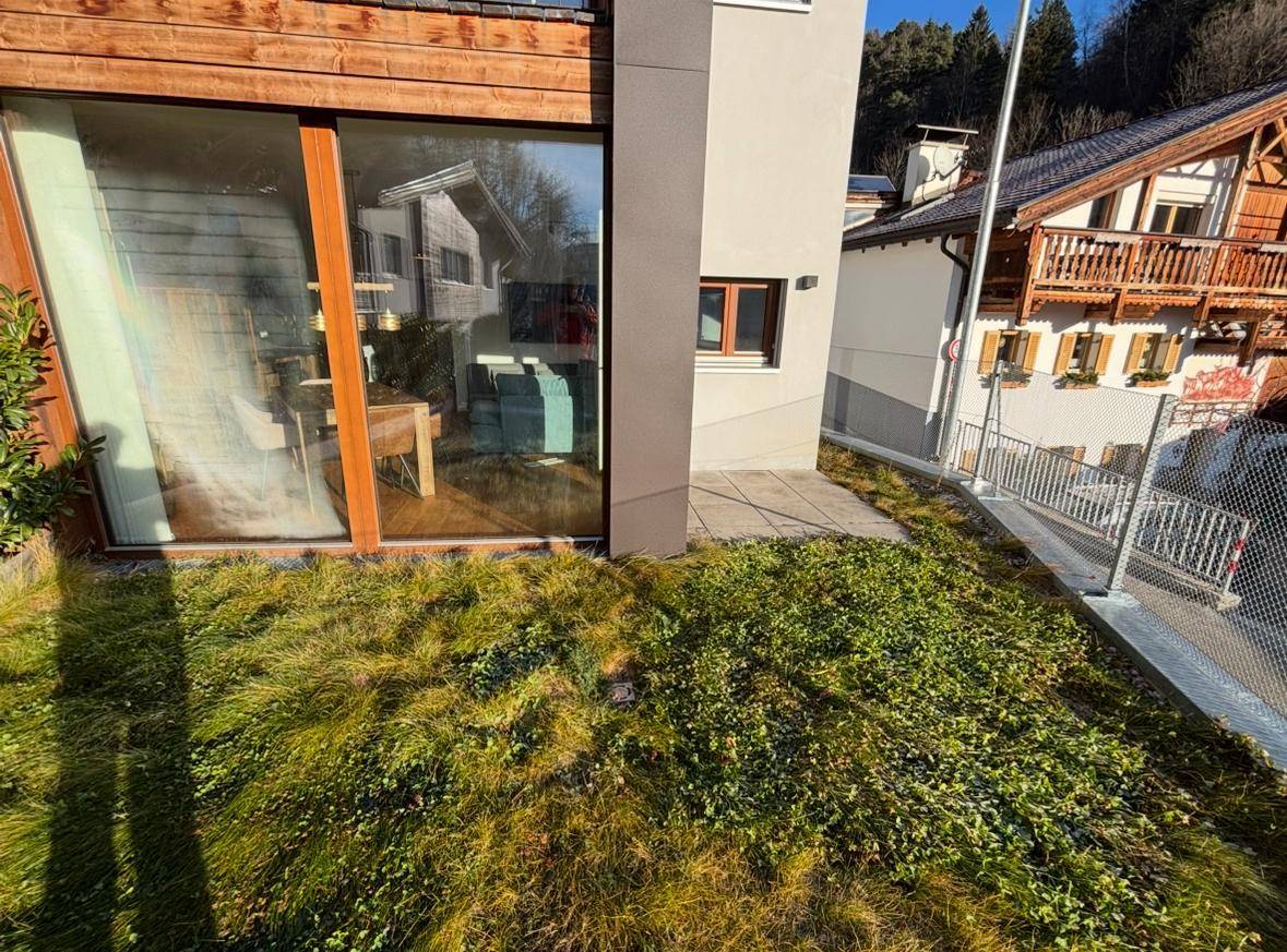 Außenansicht des Gebäudes mit großen Fenstern, einer kleinen gepflasterten Fläche und einem grünen Rasenbereich.
