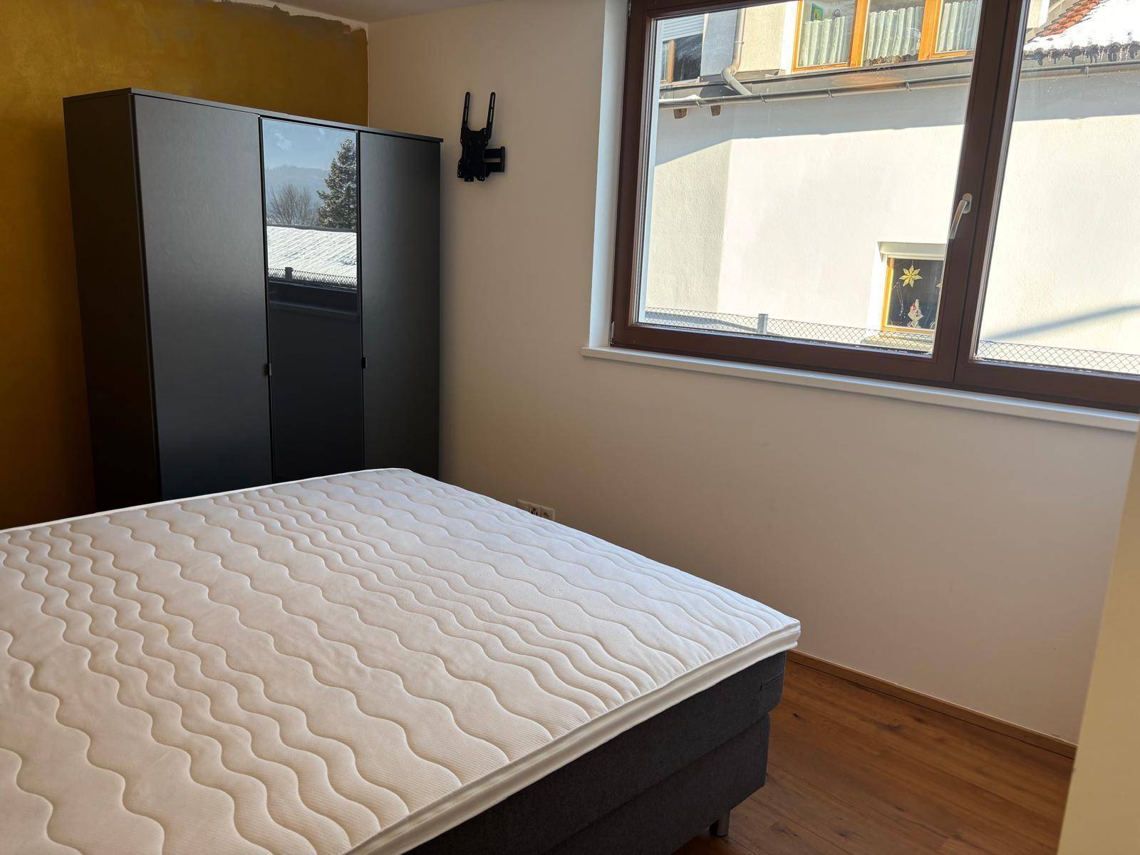 Schlafzimmer mit Bett, dunklem Kleiderschrank und Fenster, das Tageslicht in den Raum lässt.