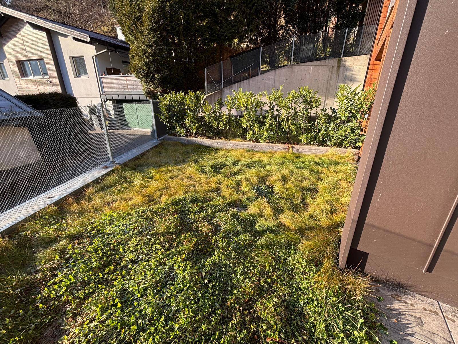 Grüner Gartenbereich neben dem Gebäude mit Zaun und Hecken im Hintergrund unter strahlendem Sonnenschein.
