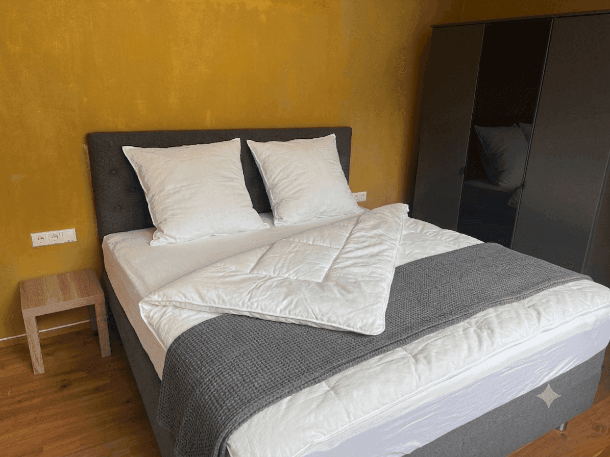 Schlafzimmer mit Doppelbett, Nachttisch und dunklem Kleiderschrank, akzentuiert durch eine gelbe Wand.