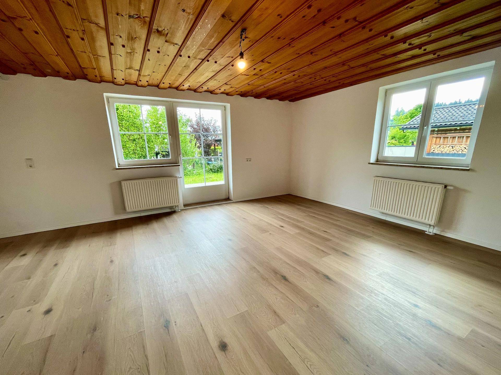 Geräumiges Zimmer mit Holzbalkendecke, hellem Holzboden und Zugang zum Garten durch eine Fenstertür.