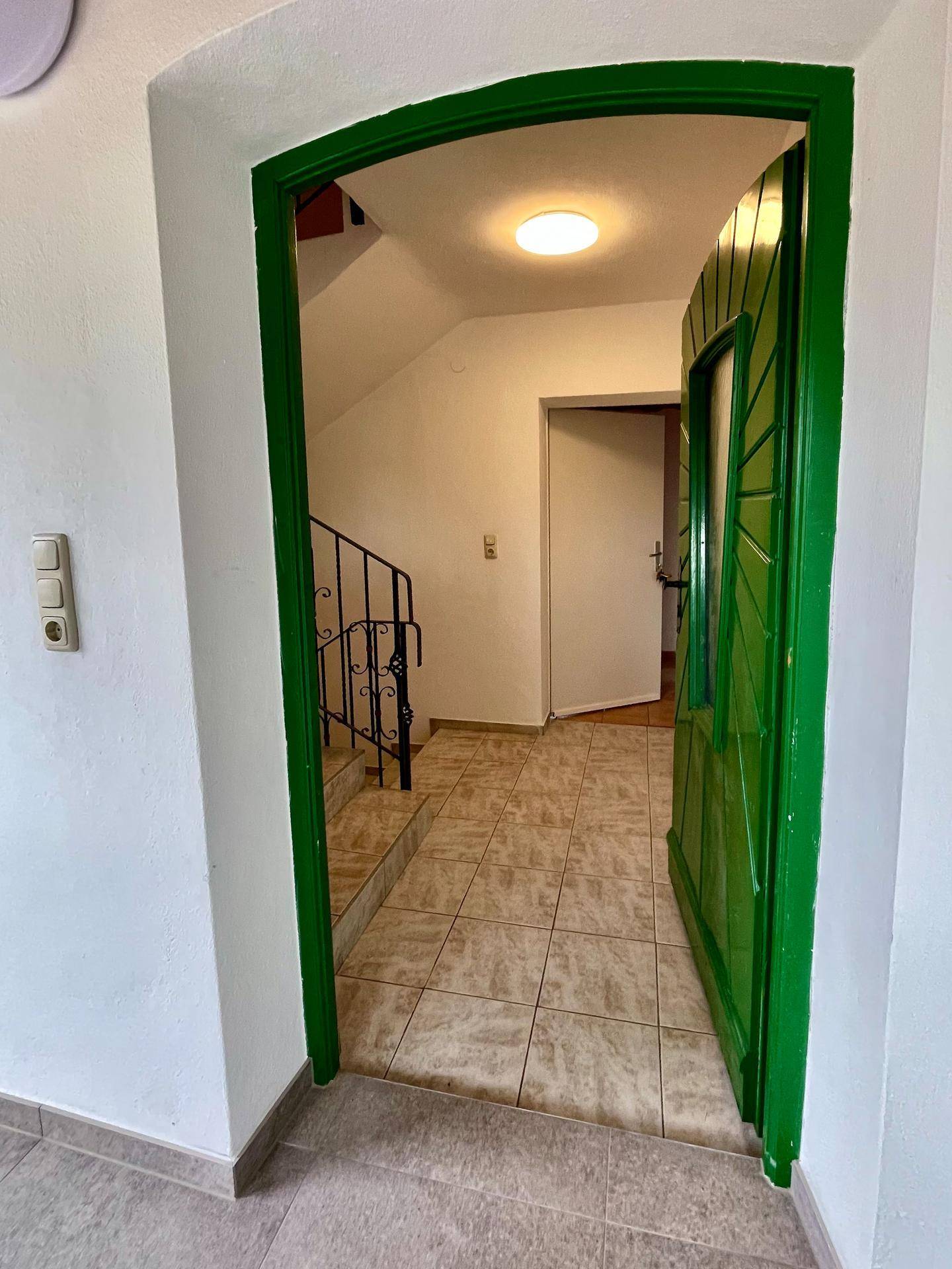 Eingangsbereich mit grüner Tür, gefliestem Boden und schmiedeeisernem Geländer zur Treppe.