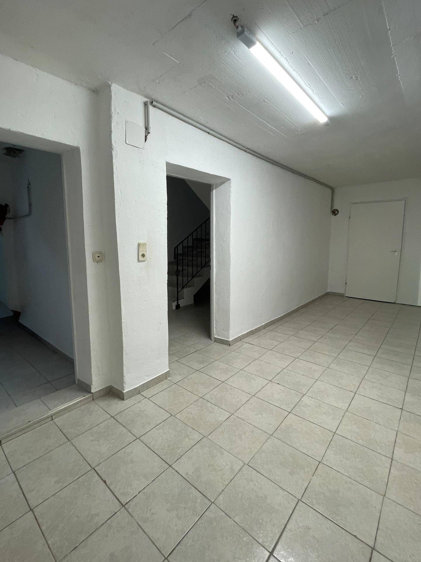 Geräumiger Kellerraum mit Fliesenboden und Zugang zu einem weiteren Bereich über eine Treppe.