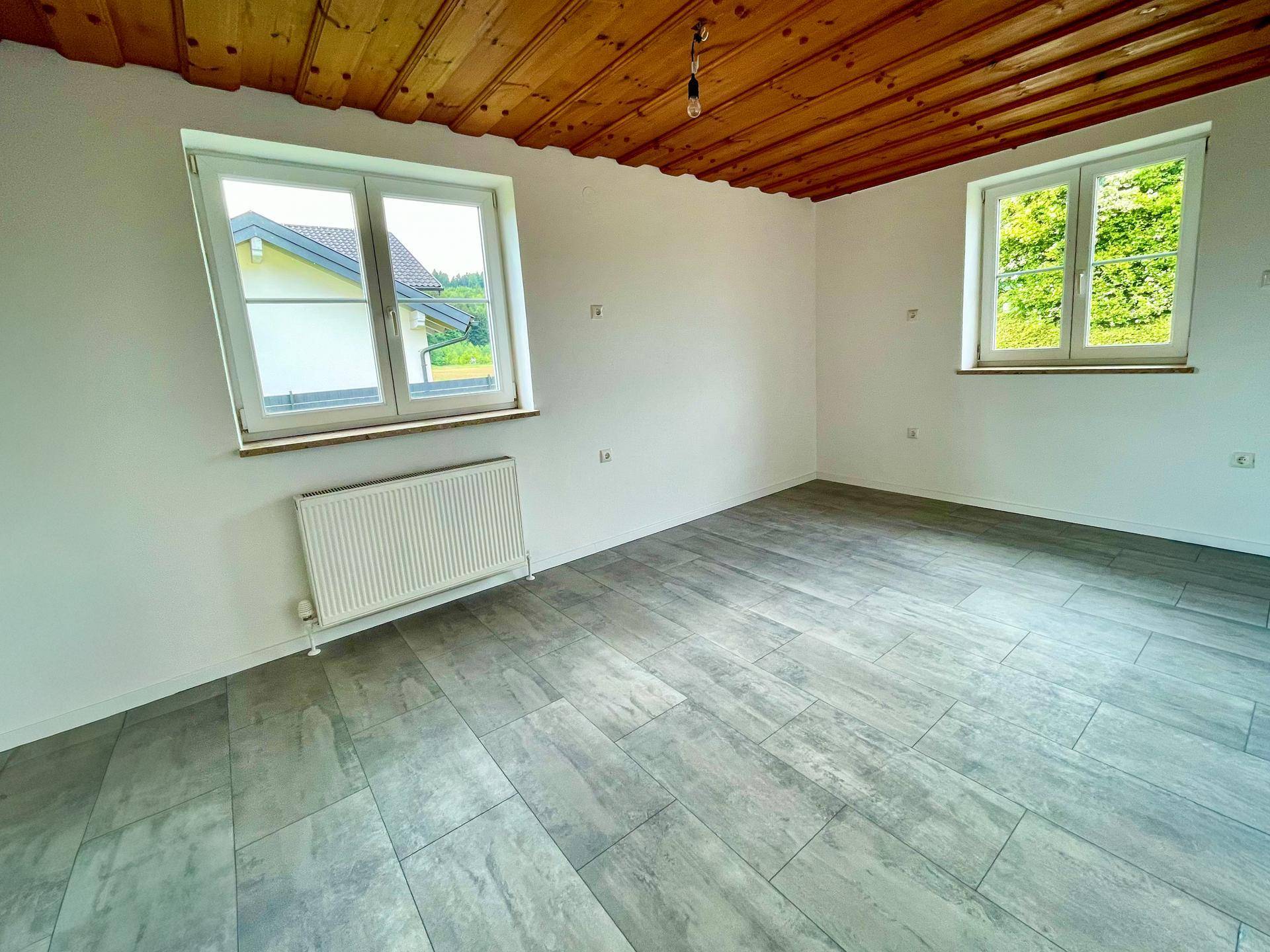Helles Zimmer mit Holzdecke, grauen Fliesen und zwei Fenstern, die den Blick ins Grüne freigeben.