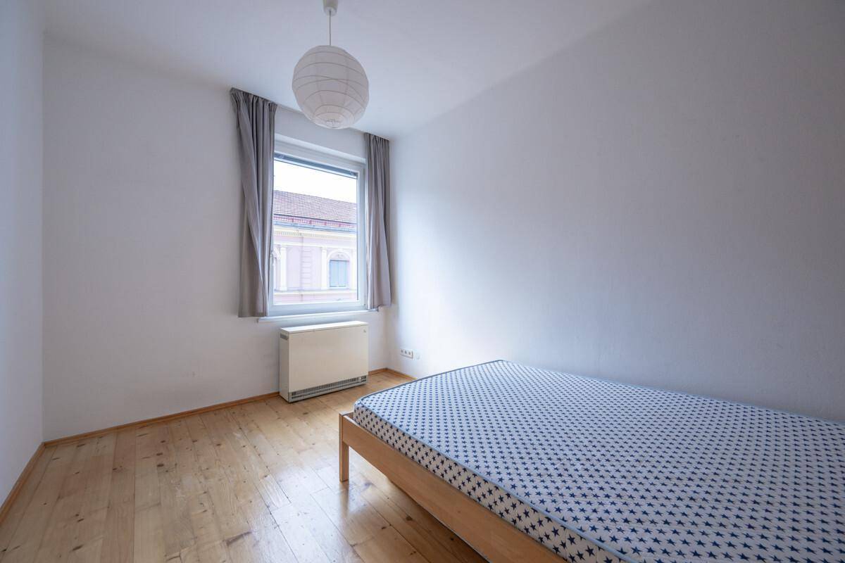 Gemütliches Schlafzimmer mit Holzboden, einem einfachen Bett und einem Fenster mit Vorhängen.