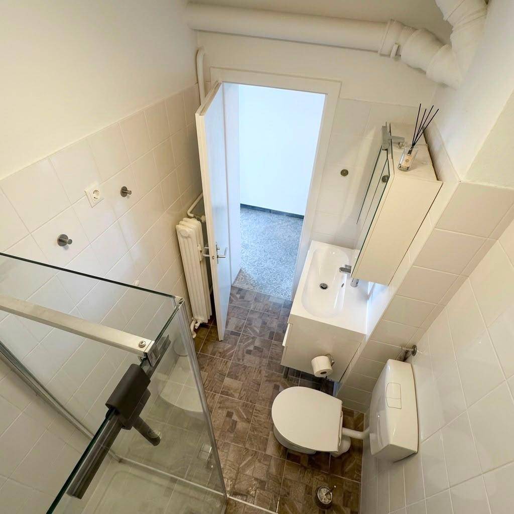Kleines Badezimmer mit Dusche, Toilette, Waschbecken und Heizkörper.