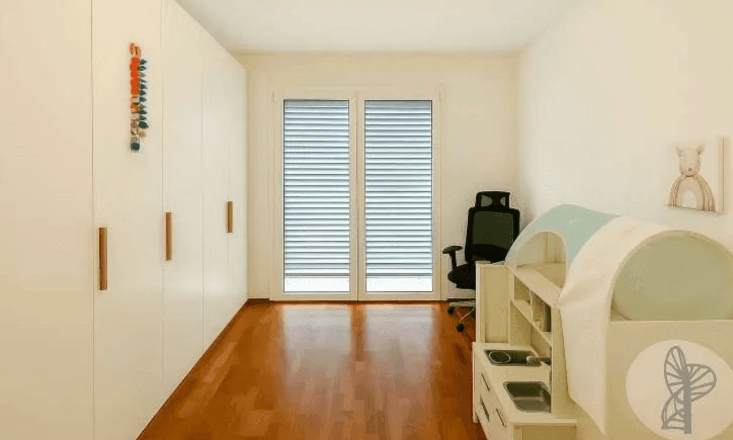 Helles Kinderzimmer mit weißen Einbauschränken, Holzboden und Fenster mit Jalousien.