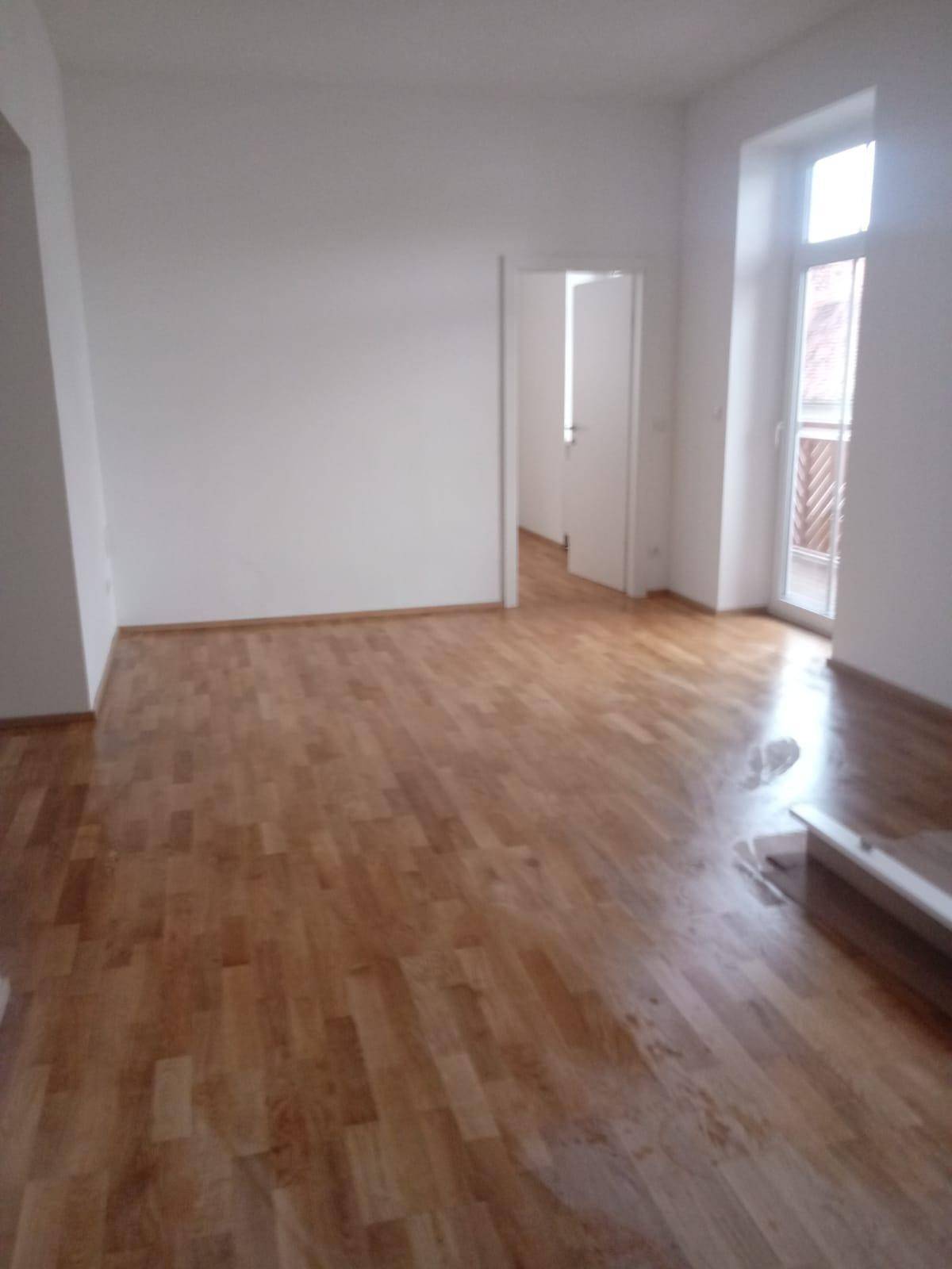Geräumiges Wohnzimmer mit glänzendem Holzparkettboden und Zugang zu einem französischen Balkon.