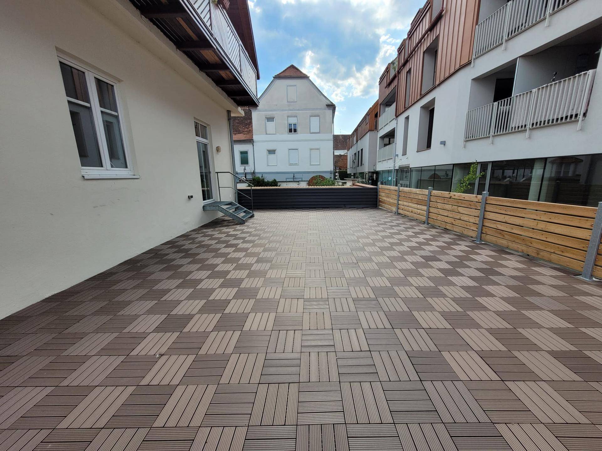 Große, geflieste Terrasse mit Holzzaun und Blick auf umliegende Gebäude.
