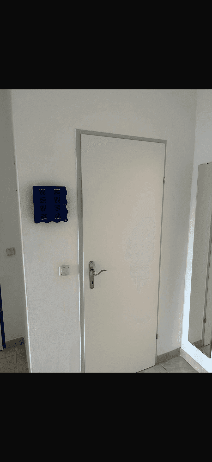 Flur mit weißer Tür, Fliesenboden und einem Wandspiegel, der den Raum erweitert.