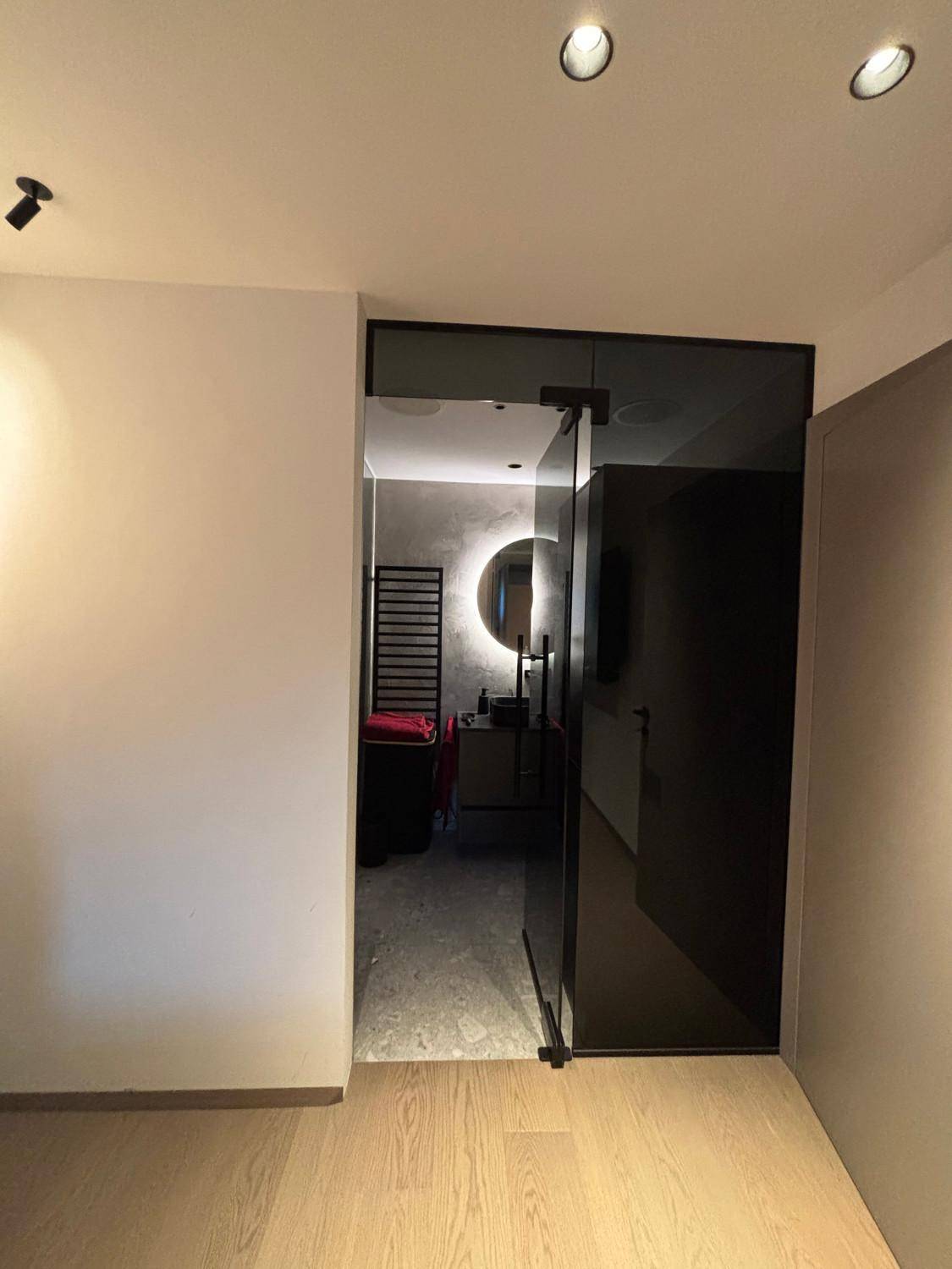 Flur mit Holzboden und Blick durch eine dunkle Glastür in ein modernes Badezimmer.