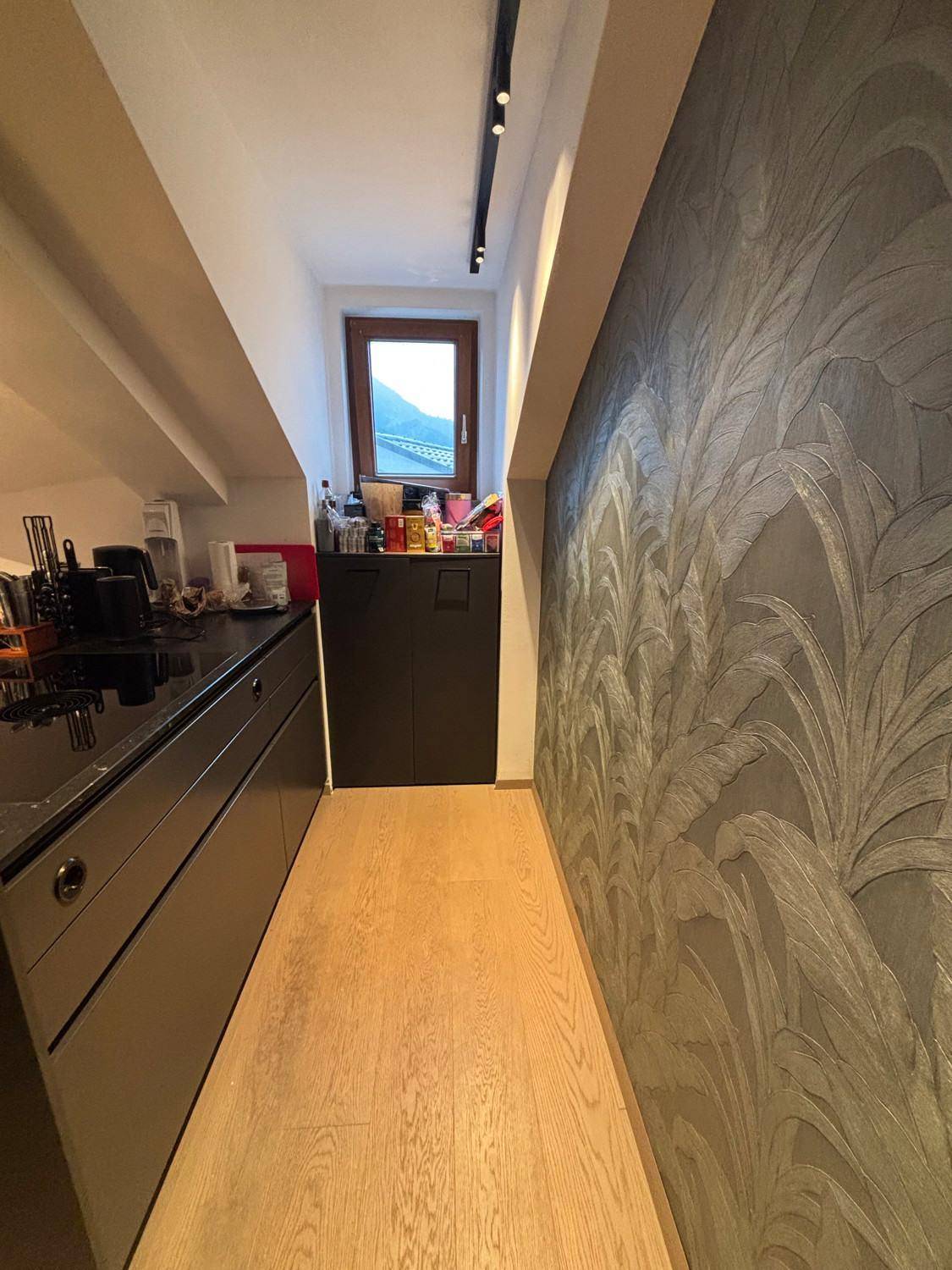 Schmaler Küchenbereich mit dunklen Arbeitsplatten, Schränken und gemusterter Tapete.