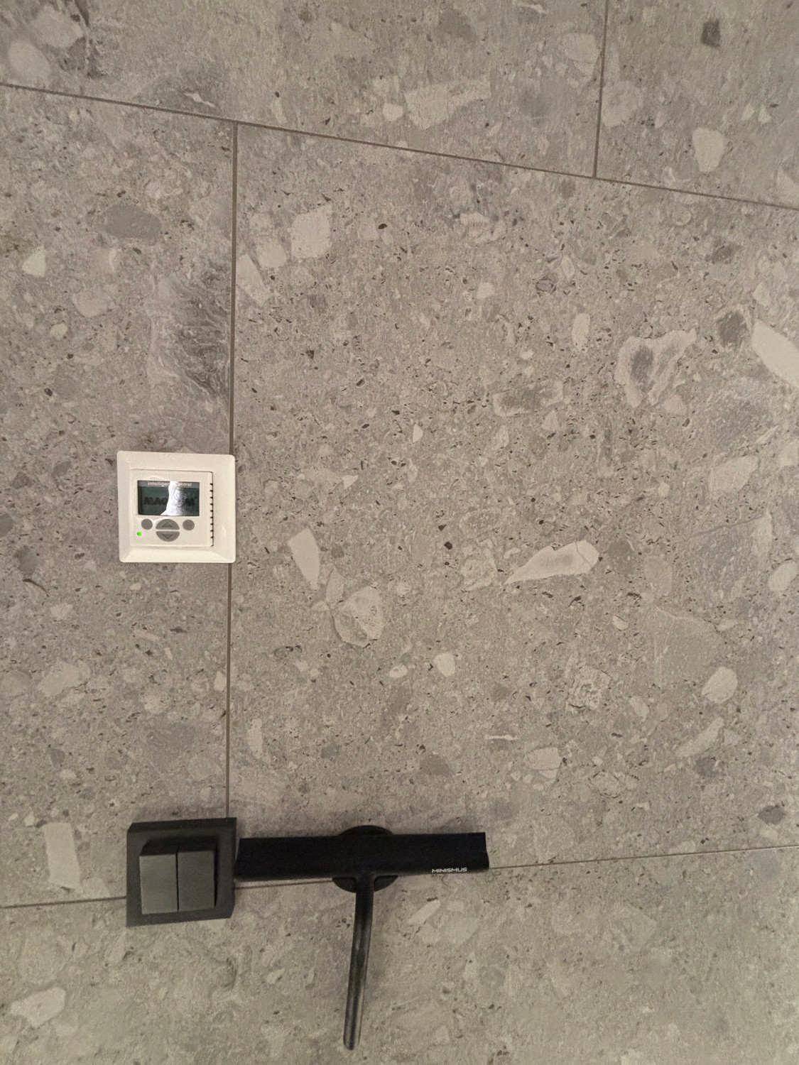 Nahaufnahme einer grauen Fliesenwand mit Thermostat und modernem Lichtschalter.