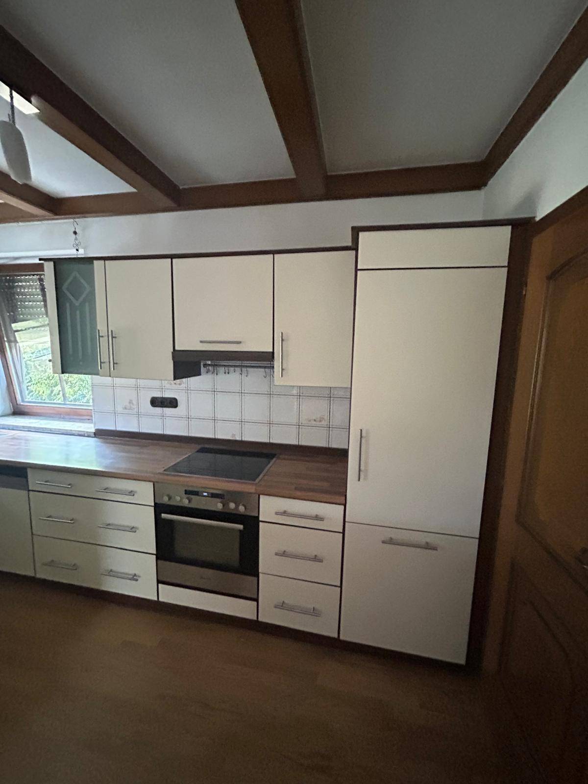 Einbauküche mit hellen Fronten, integriertem Backofen und Kühlschrank, unter Holzbalken.