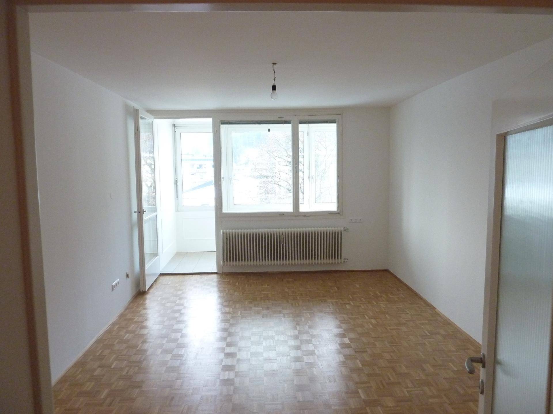 Leeres Zimmer mit Parkettboden, großen Fenstern und Zugang zu einem verglasten Balkon.