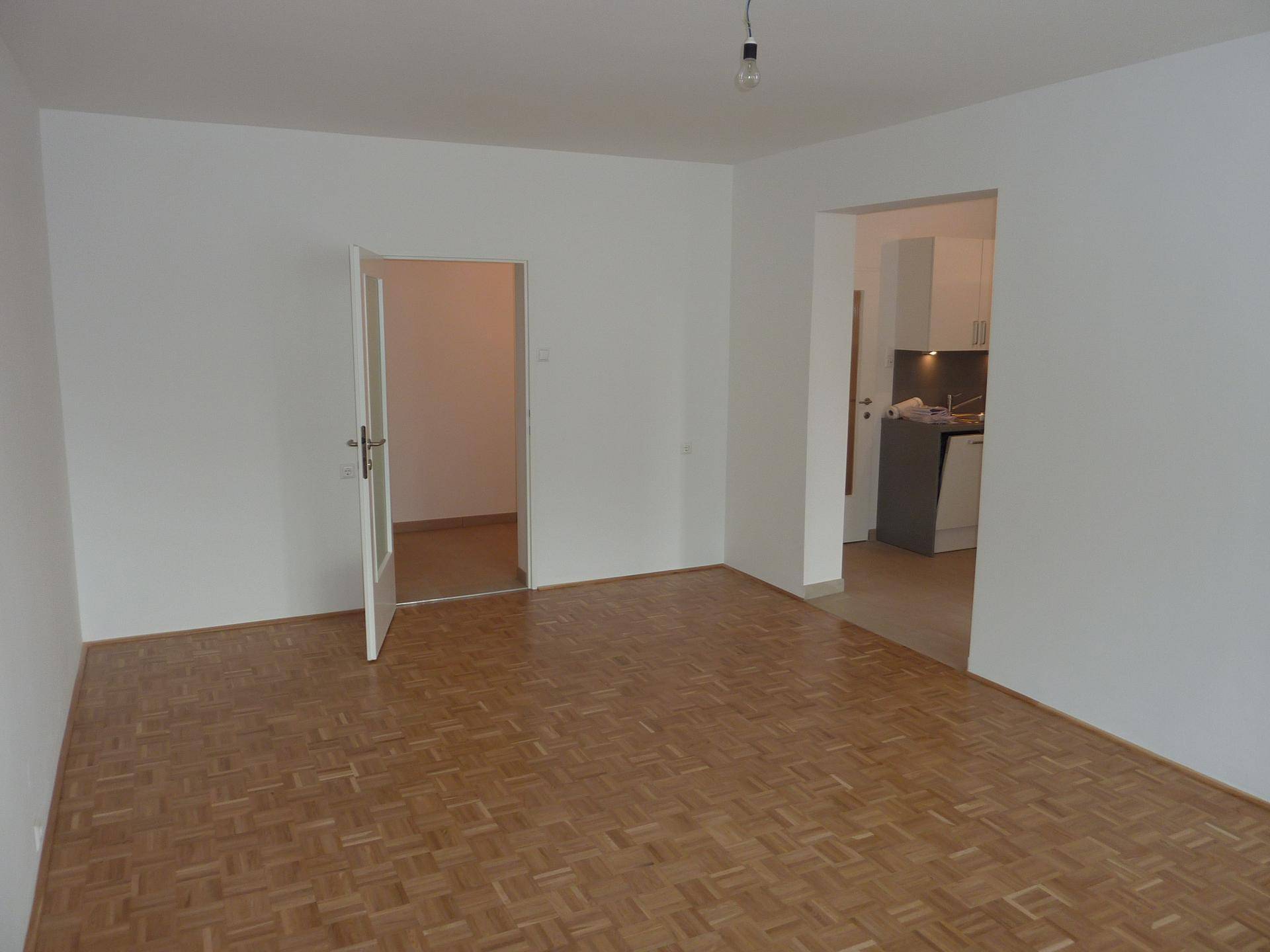 Wohnzimmer mit Parkettboden und weiß gestrichenen Wänden, offen zur angrenzenden Küche.