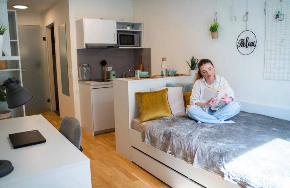 Junge Frau liest auf dem Bett in einem hellen Studio-Apartment mit integrierter Küchenzeile.