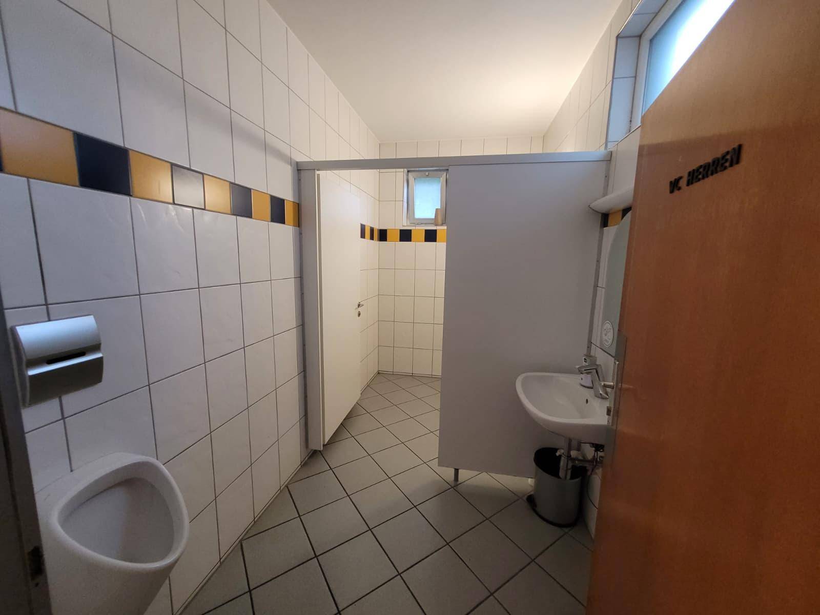 Herren-Waschraum mit Urinal, Waschbecken und gefliesten Wänden, gut beleuchtet.