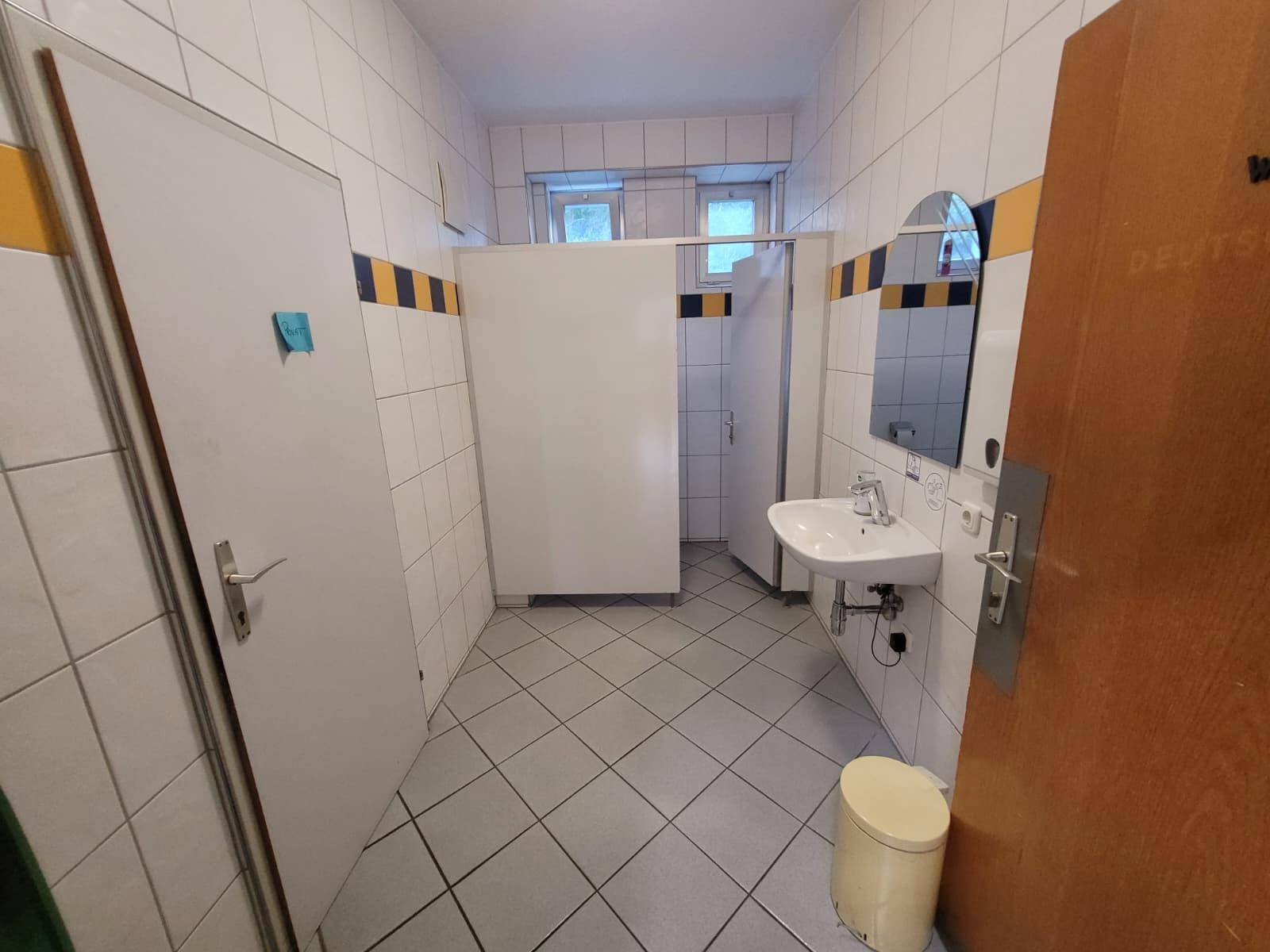 Gefliester Waschraum mit Waschbecken und Toilettenkabinen, ausgestattet mit einem Fenster.