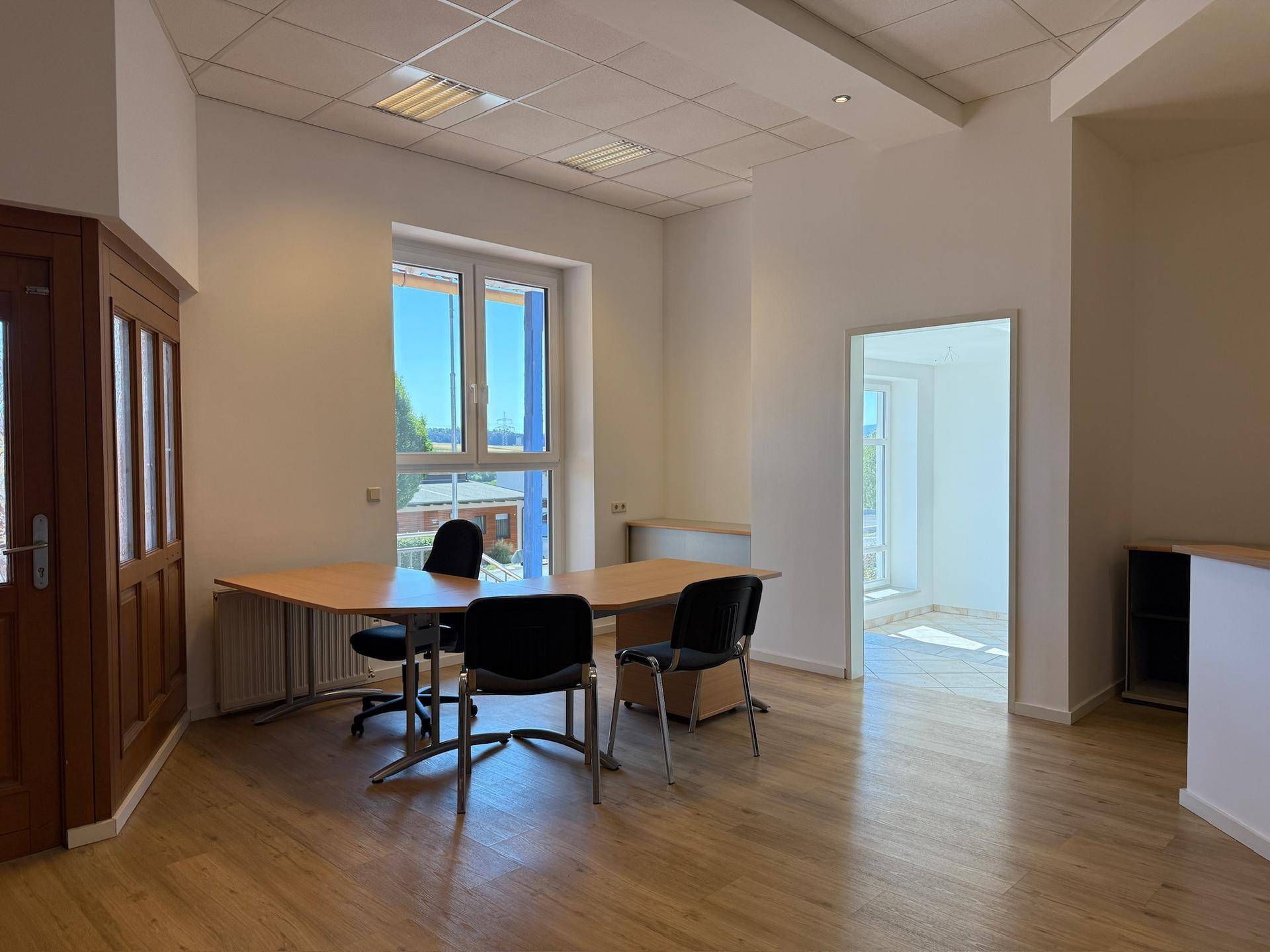 Geräumiges Büro mit Holztür, Laminatboden und großen Fenstern, die viel Tageslicht hereinlassen.