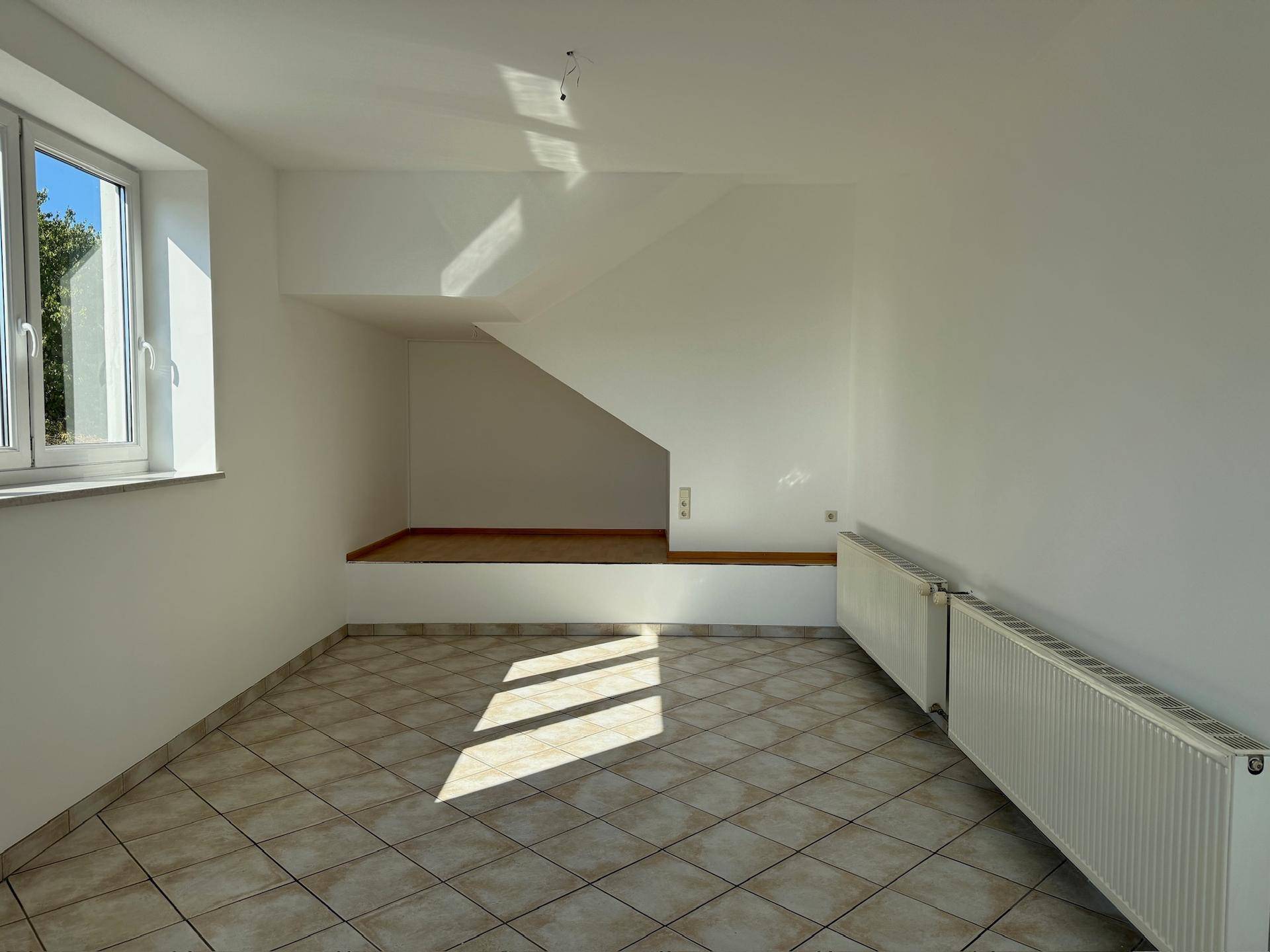 Leerer Raum mit Fliesenboden, Heizkörpern und einem Fenster, das Sonnenlicht hereinlässt.